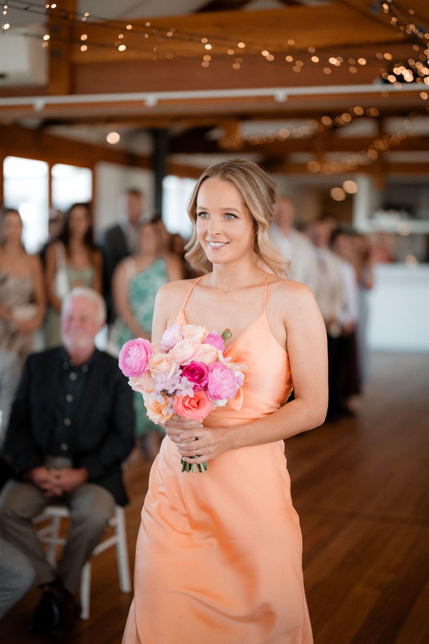 Bridesmaid in peach dress carrying pink bouquet walks down aisle at indoor wedding venue.