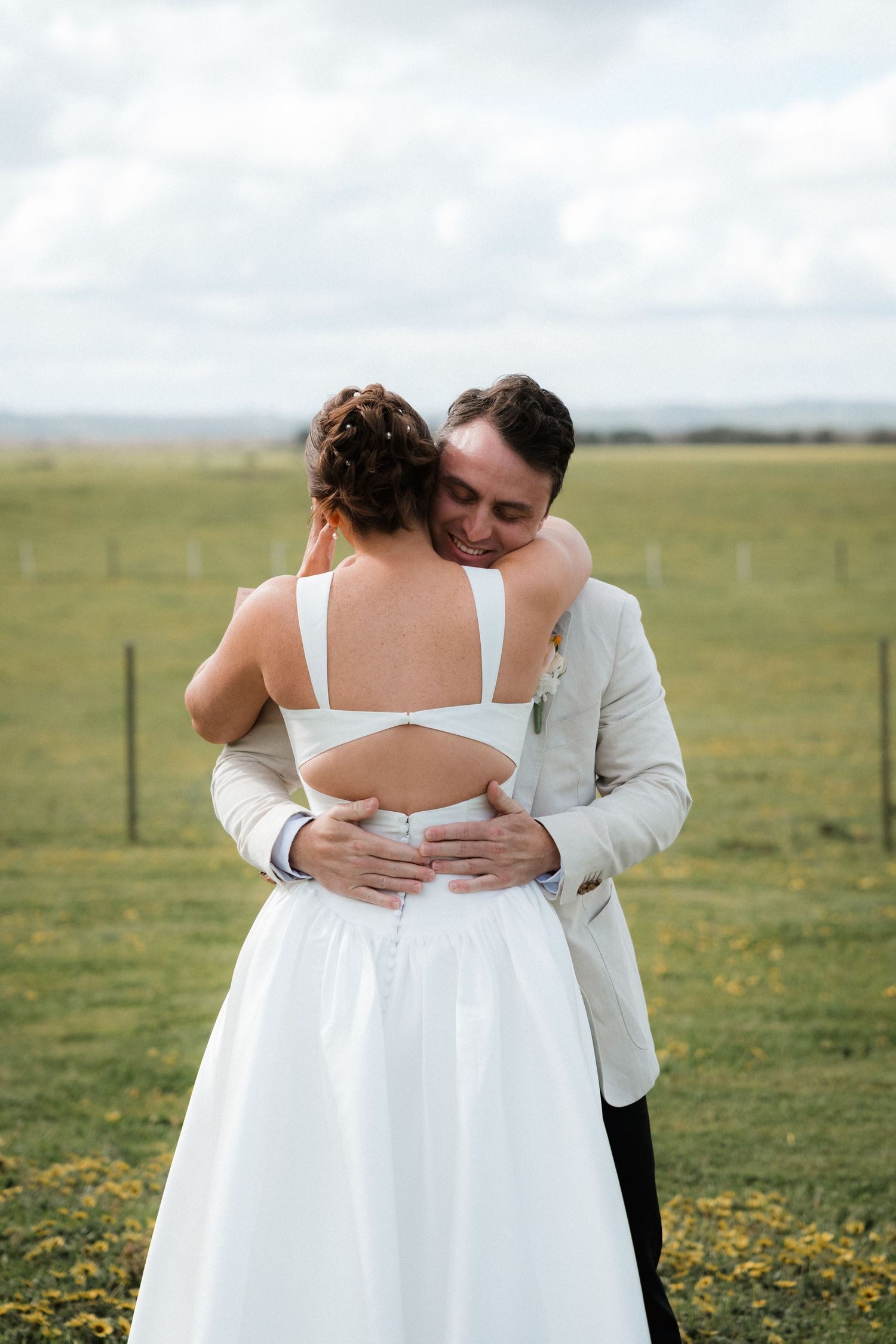 A tender embrace captured in a field of wildflowers with white wedding attire against a cloudy sky.