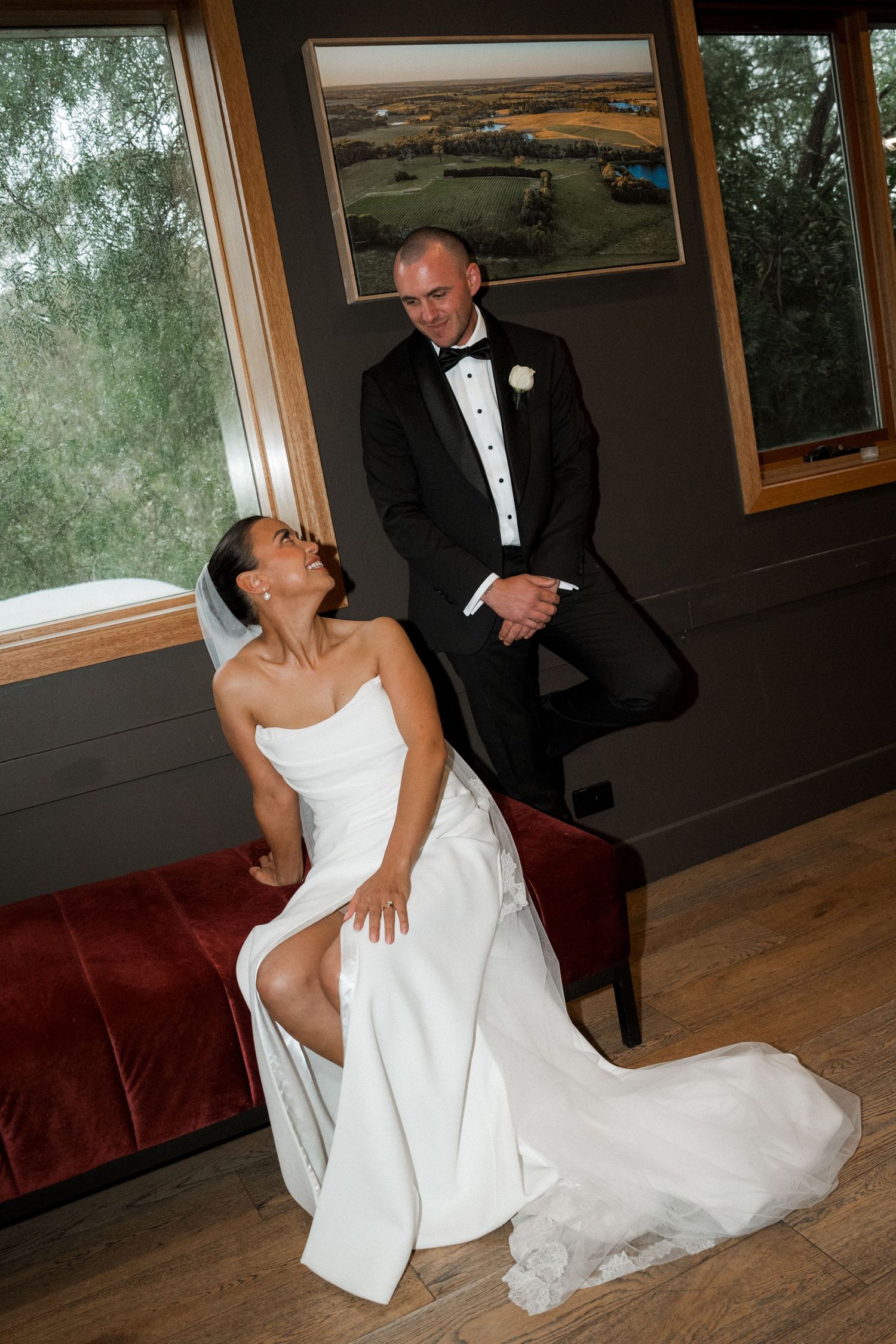 Bride in white wedding dress lounges playfully on red couch during indoor photo session.