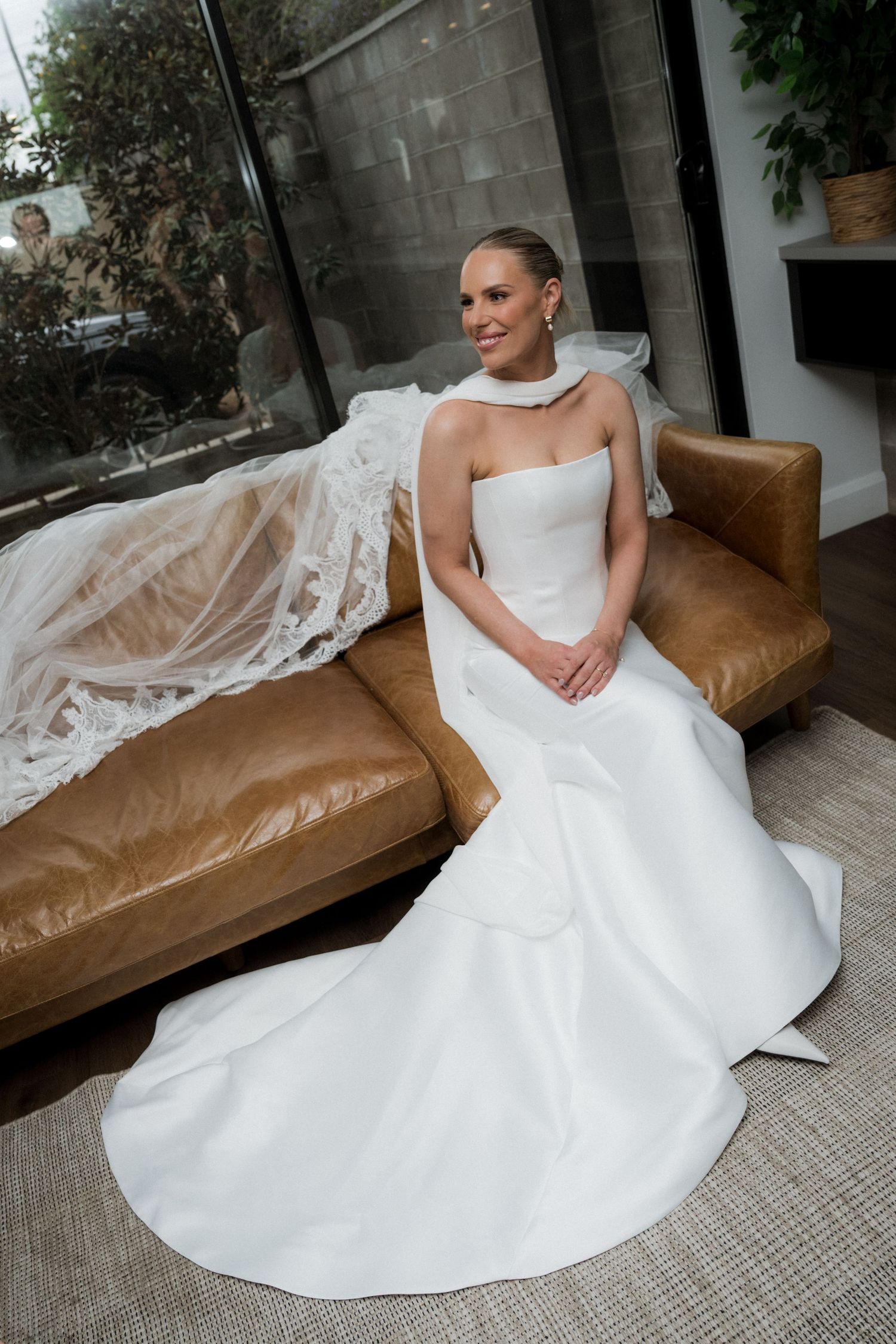 A bride in an elegant white wedding gown poses gracefully on a brown leather couch.