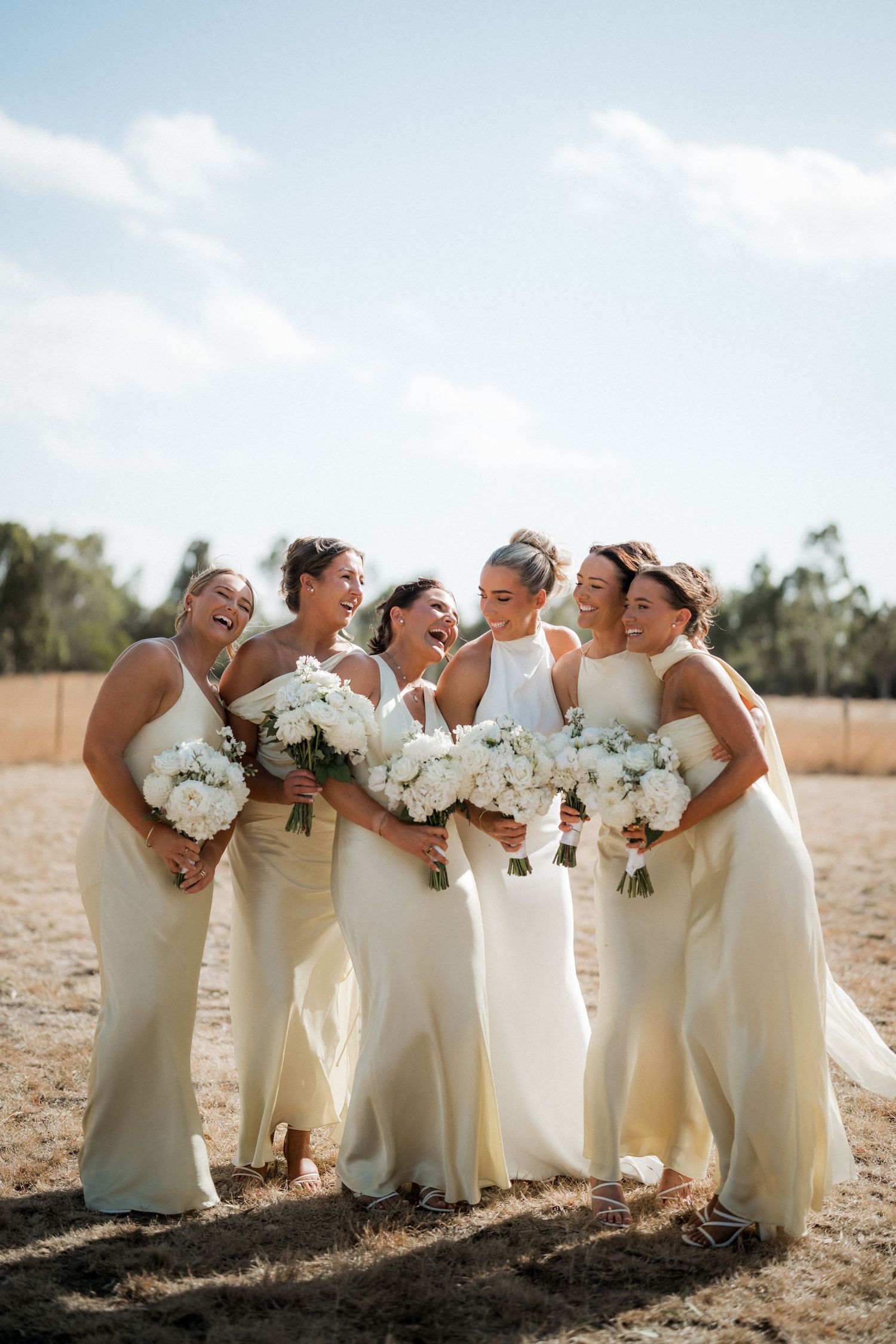 Bridesmaids in cream-colored dresses laugh together while holding white bouquets in a sunlit field.