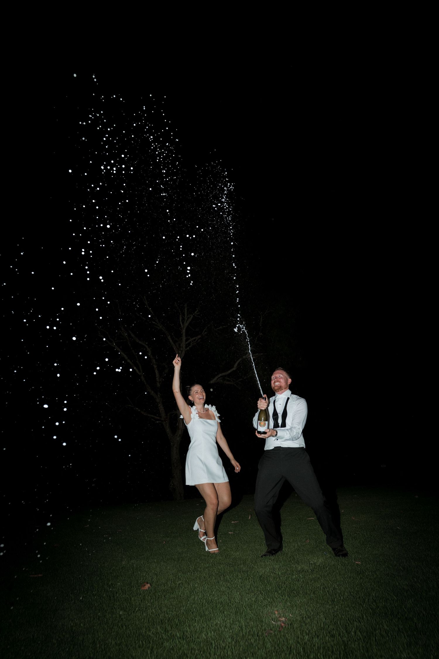 Wedding party members play with sparklers at night on a dark grassy lawn.