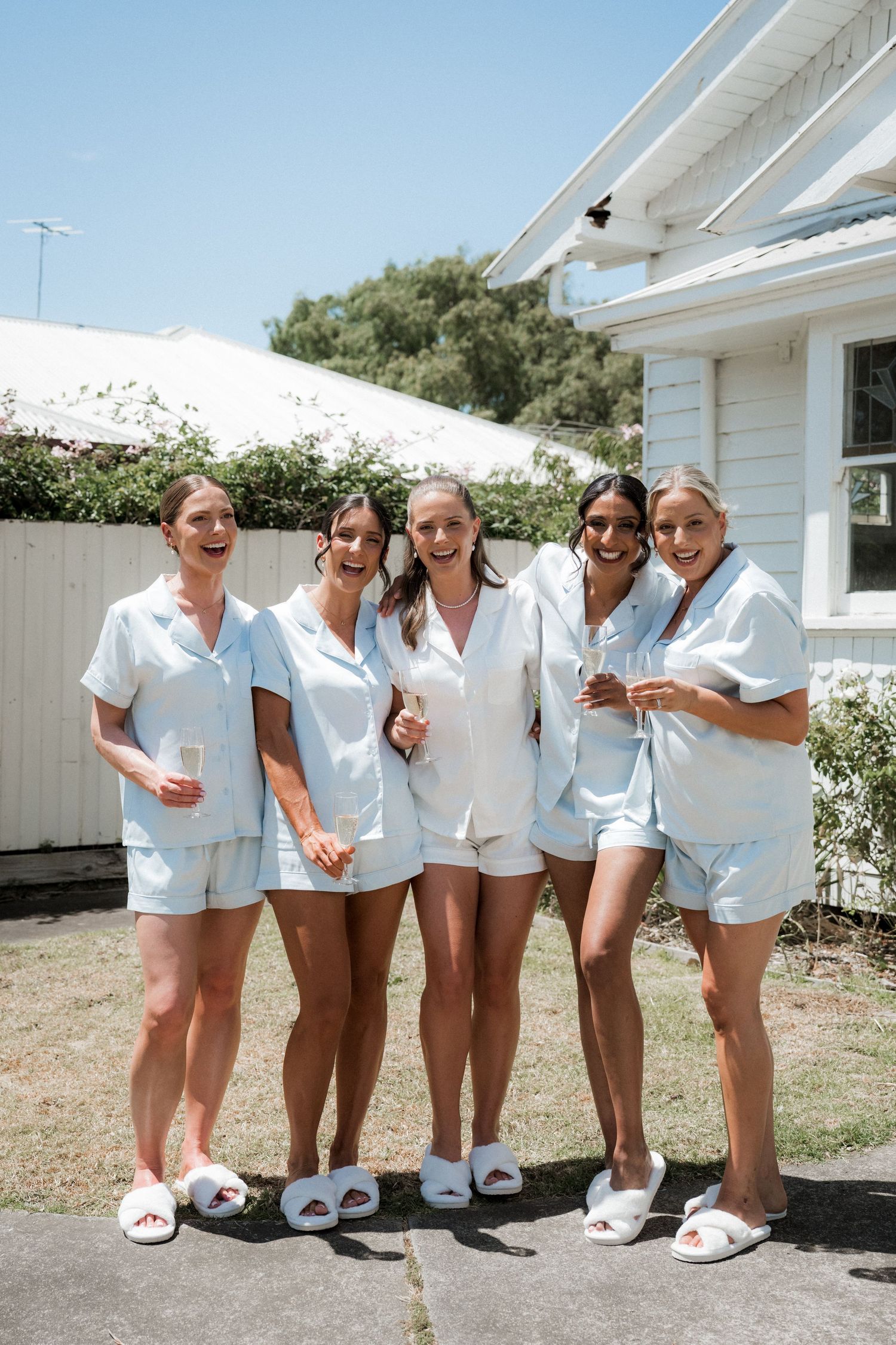 A group in matching white outfits and slippers pose playfully together outdoors against a white building.