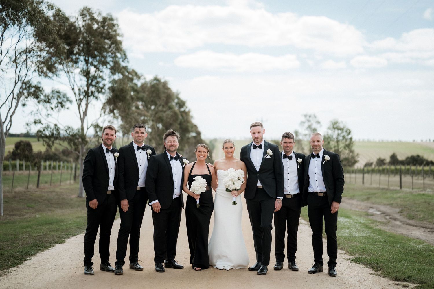 Wedding party in formal attire poses together for outdoor photos on a rural property with trees in background.