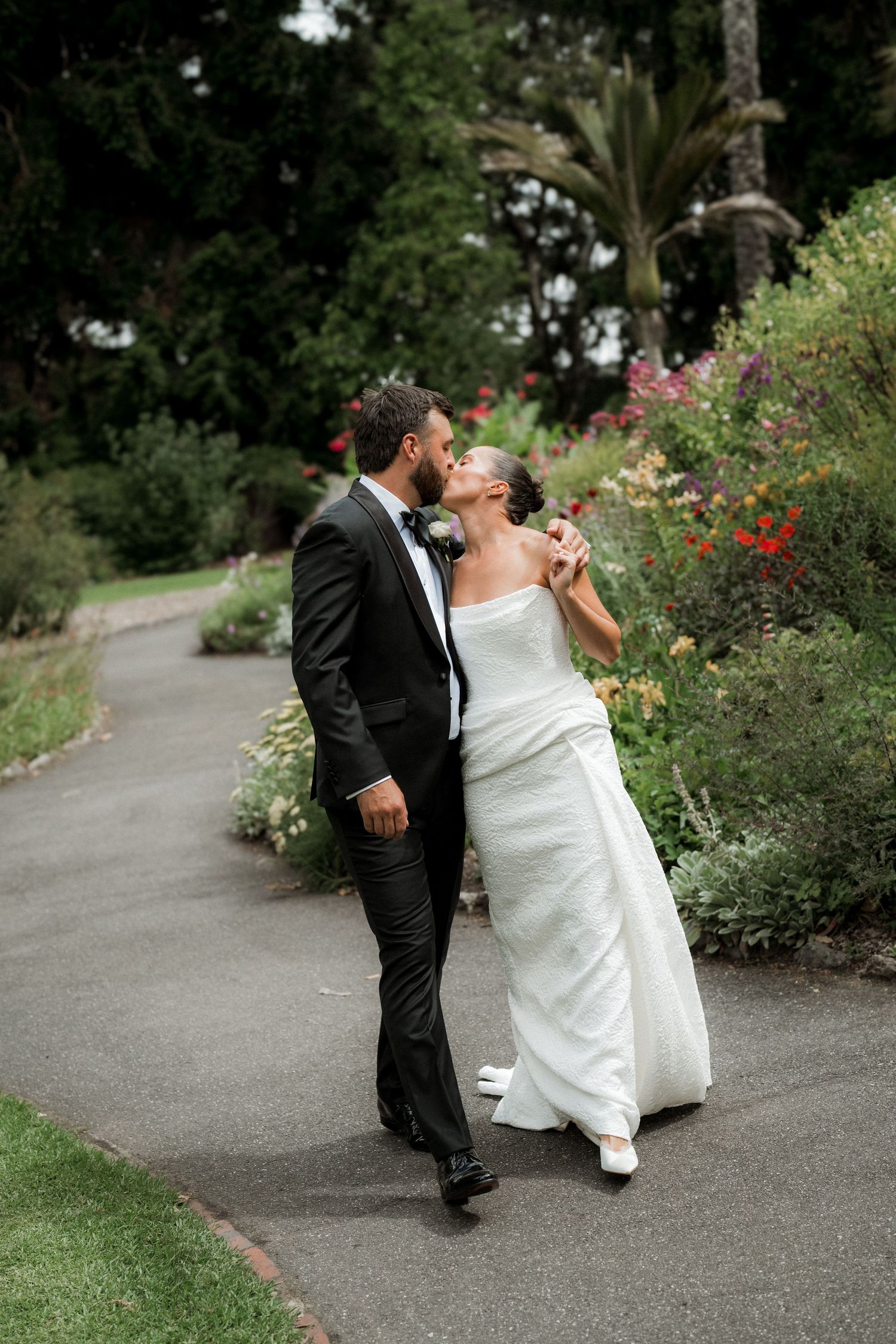 Bride and groom sharing a kiss while walking along a garden path surrounded by colorful flowers.