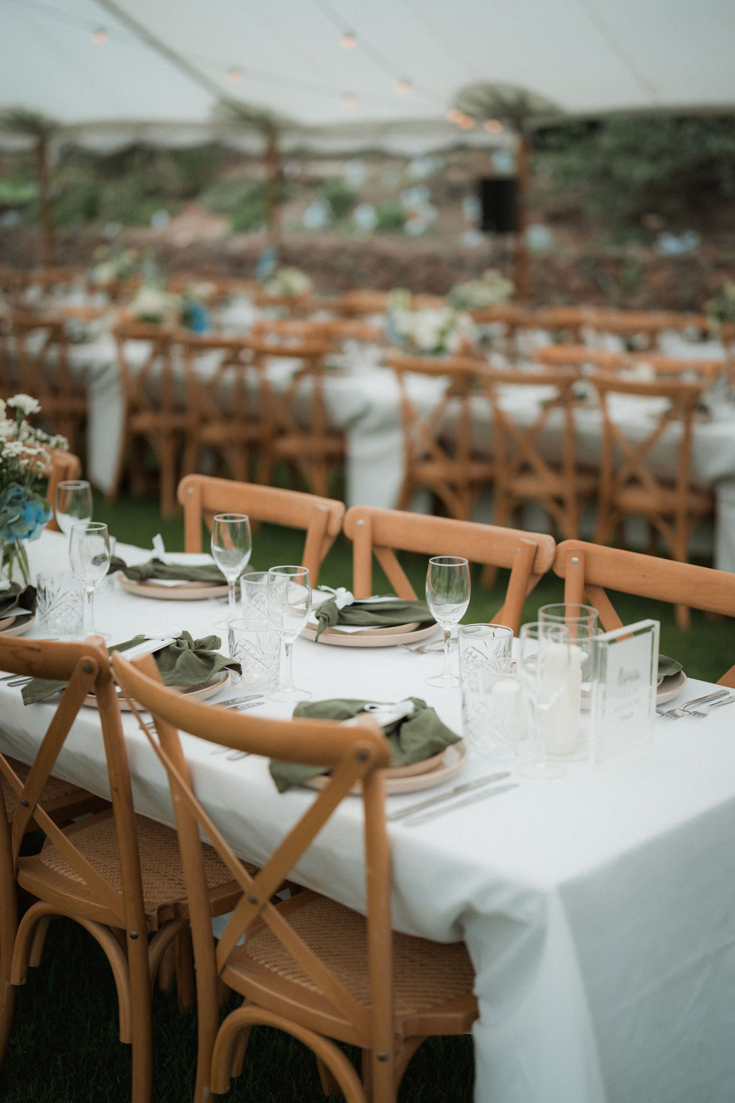 Elegant outdoor wedding reception table setting with wooden cross-back chairs and white linens.