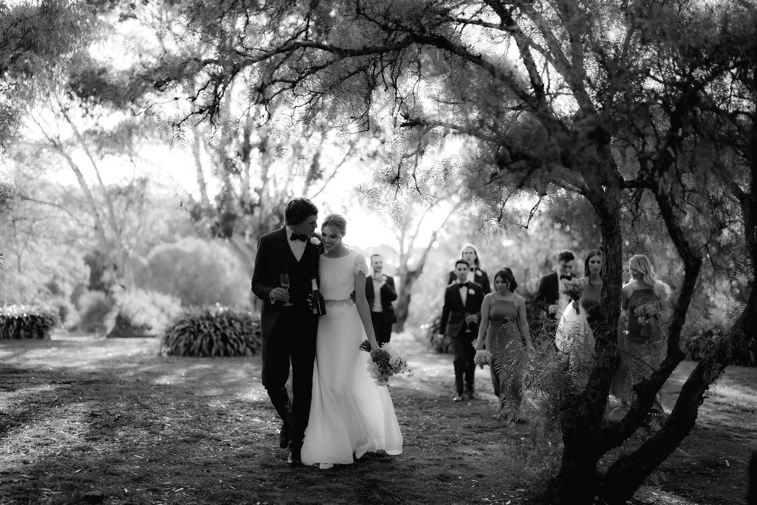 Black and white wedding photo of couple walking through tree-lined path with guests.