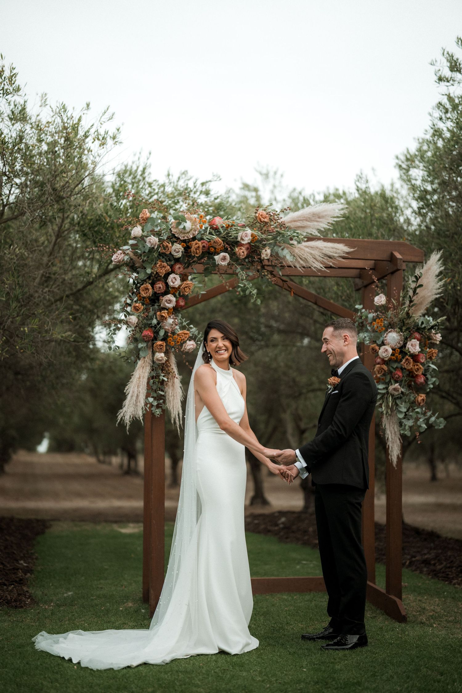 Couple holds hands under a wooden arch decorated with peach roses and pampas grass during their outdoor wedding ceremony.