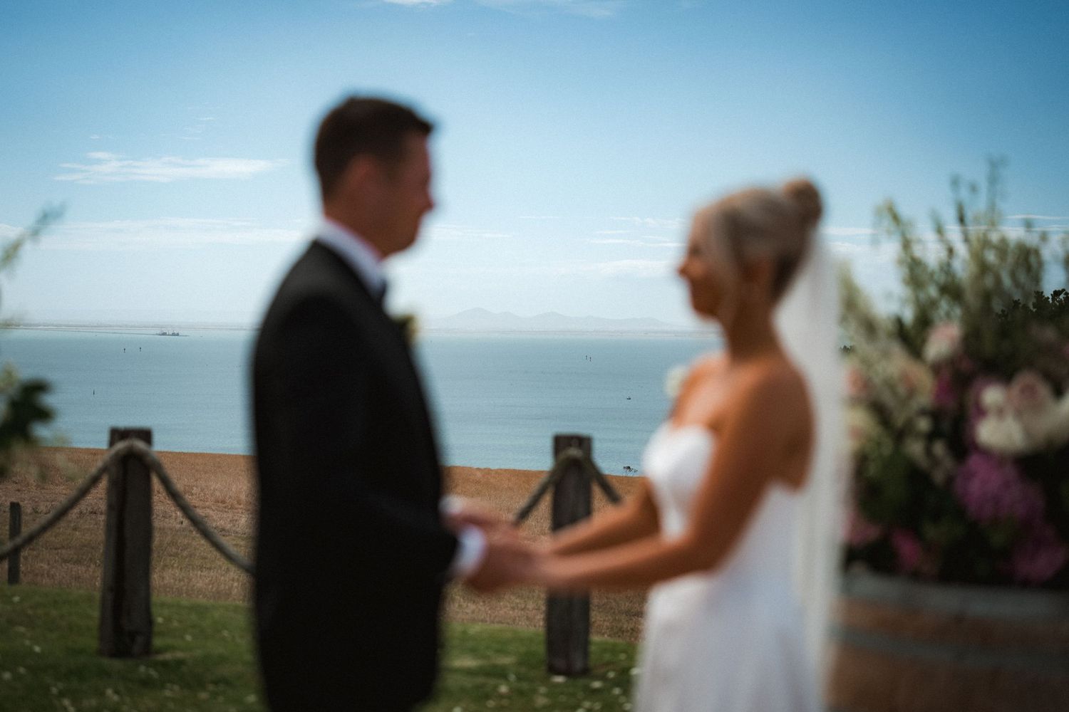 Two wedding ceremony participants exchange vows at an outdoor coastal venue with ocean views and floral decorations.