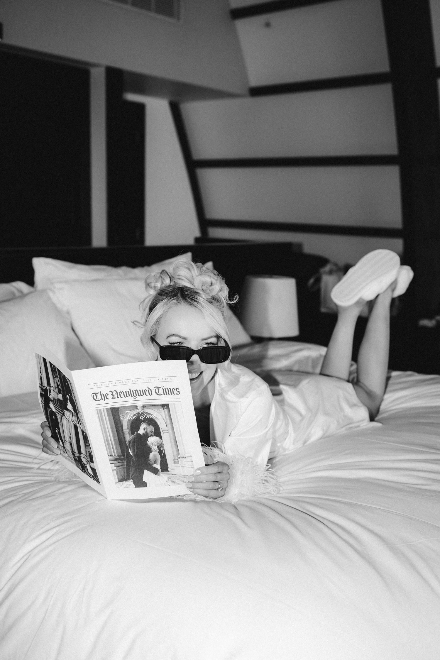 Person relaxing on white bed while reading a newspaper in black and white photography.