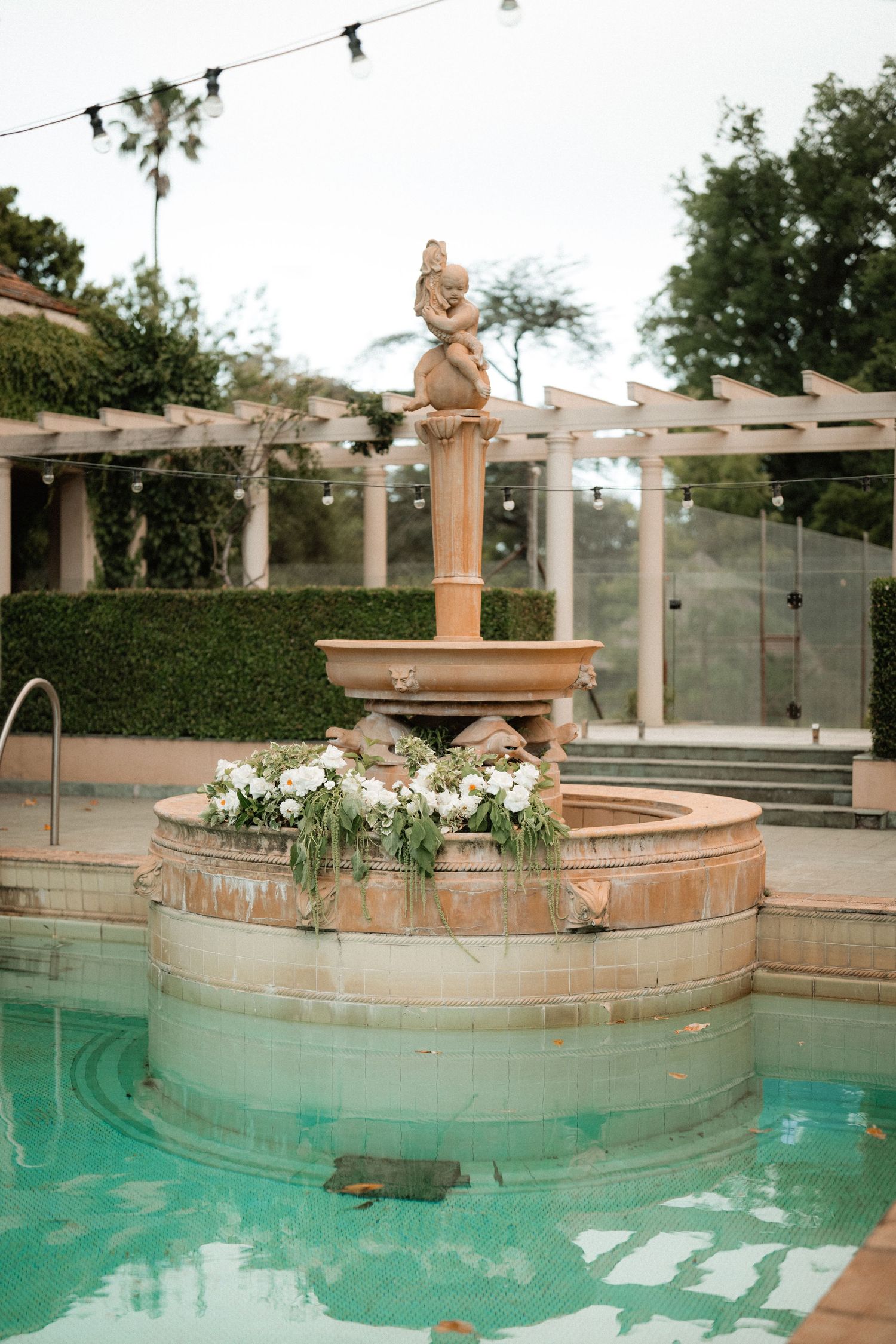 An elegant stone fountain with floral decorations sits in a turquoise pool surrounded by a Mediterranean-style pergola.