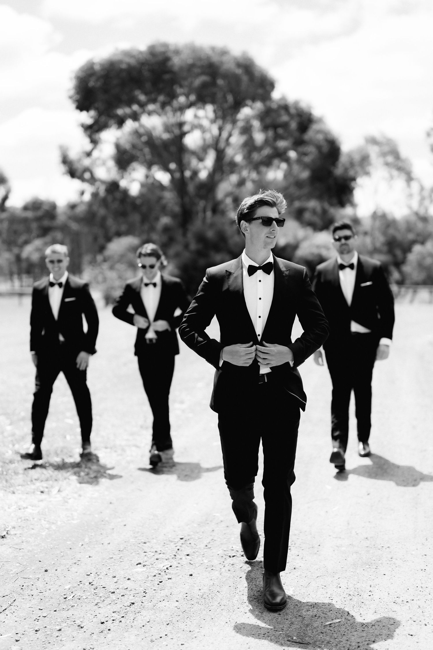 Groomsmen in black tuxedos pose confidently on a dirt path with trees in background for stylish wedding photo.