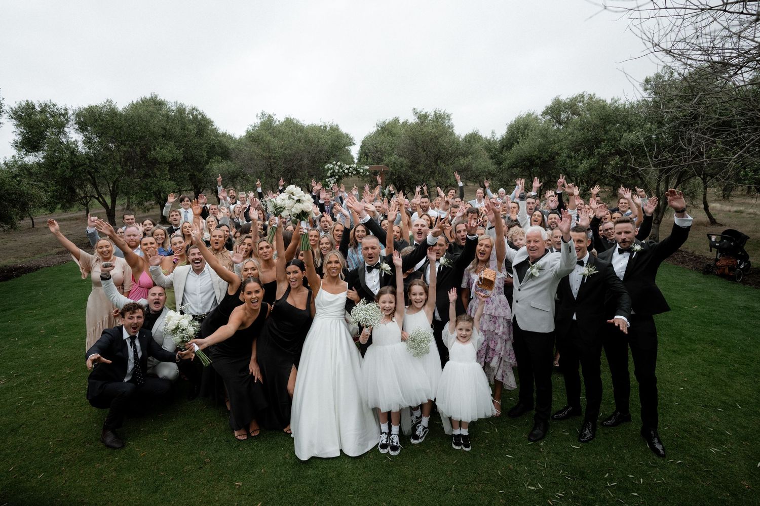 A large wedding party poses together outdoors with raised arms in celebration.