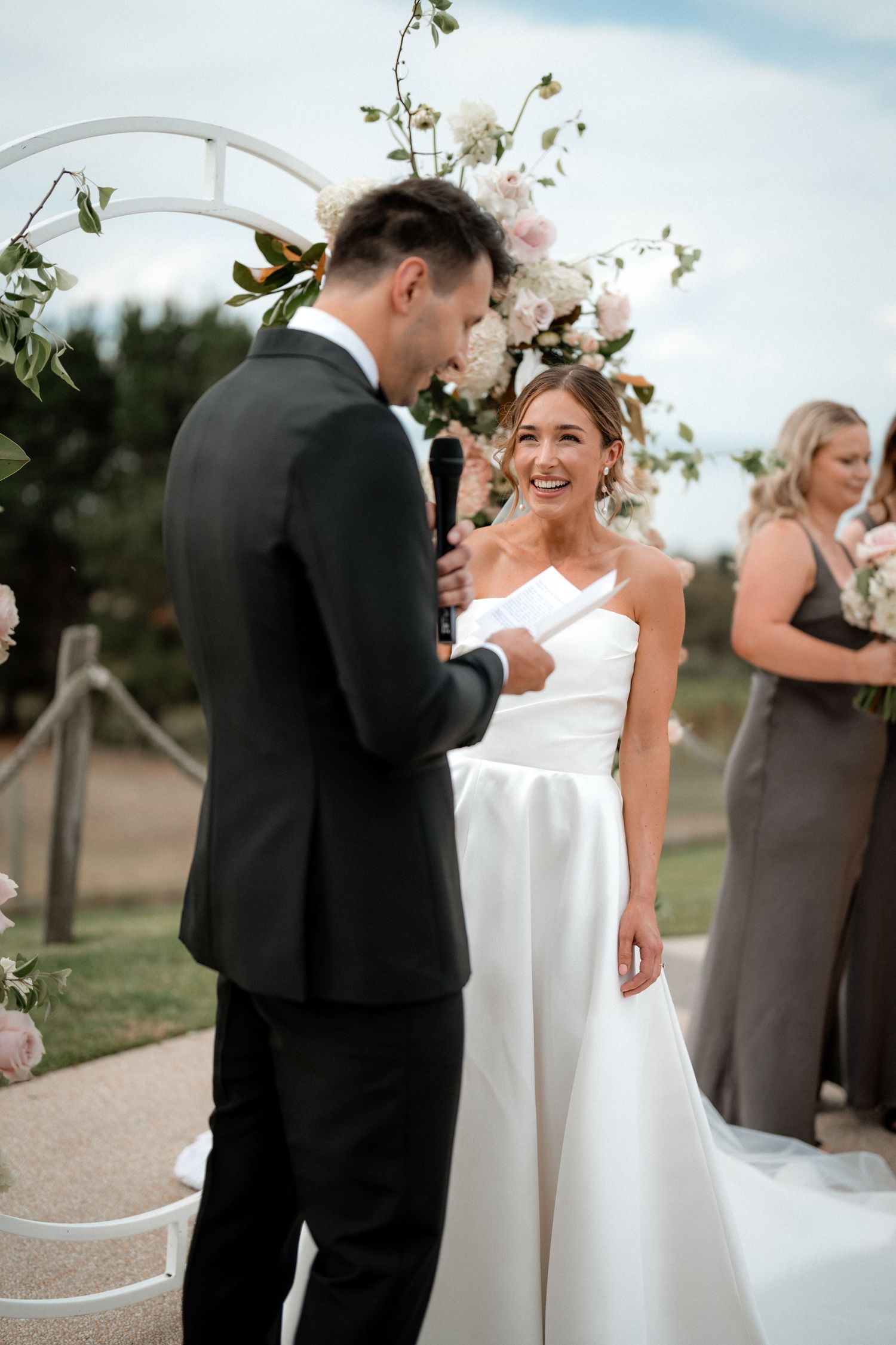 Wedding couple shares a joyful moment during their ceremony under a floral arch with guests looking on.