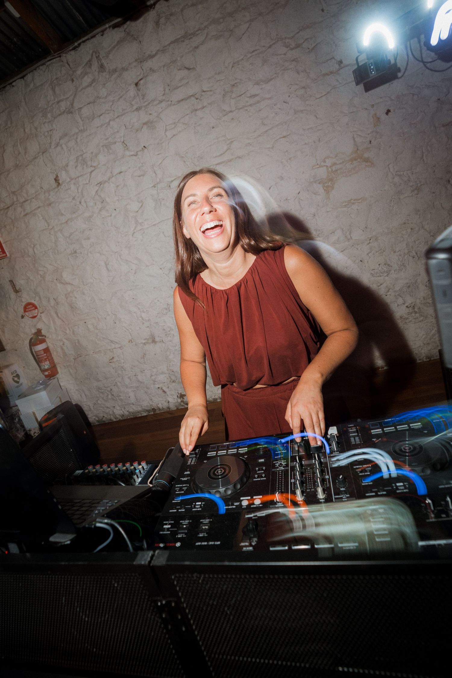 Female DJ laughs joyfully while mixing music at a wedding reception in a rustic white-walled venue.