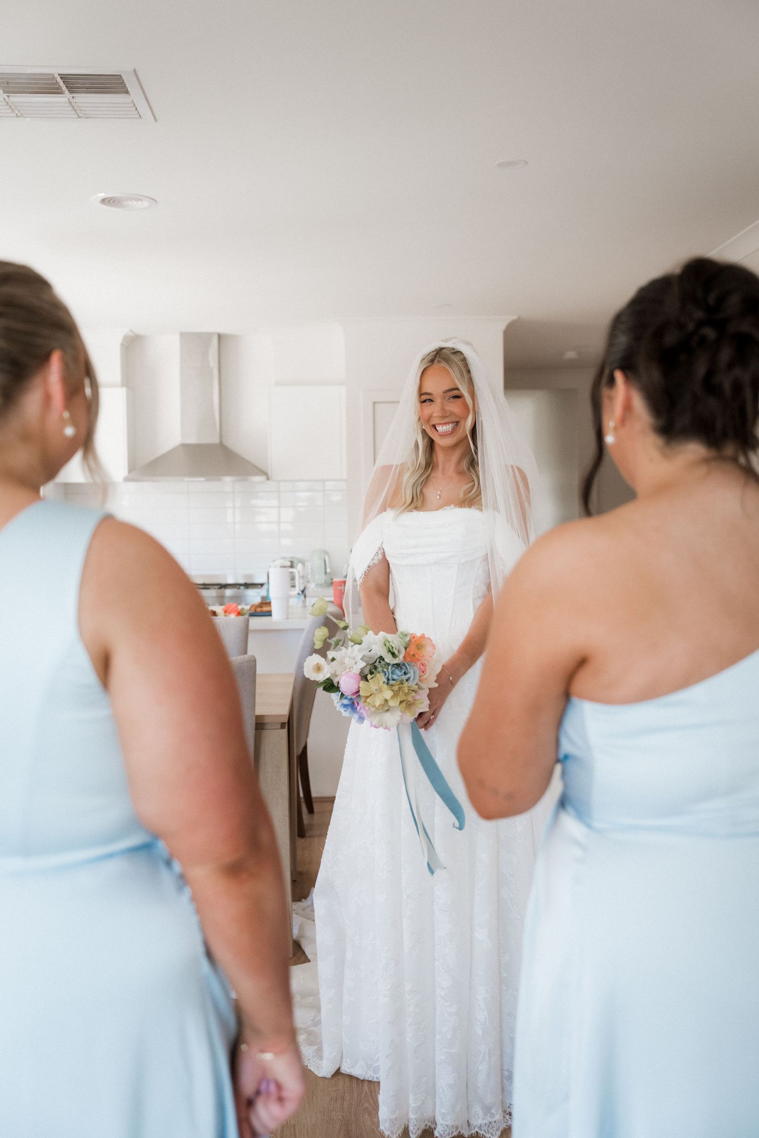 Bride in white lace gown holding floral bouquet smiles at bridesmaids in light blue dresses during wedding preparations.