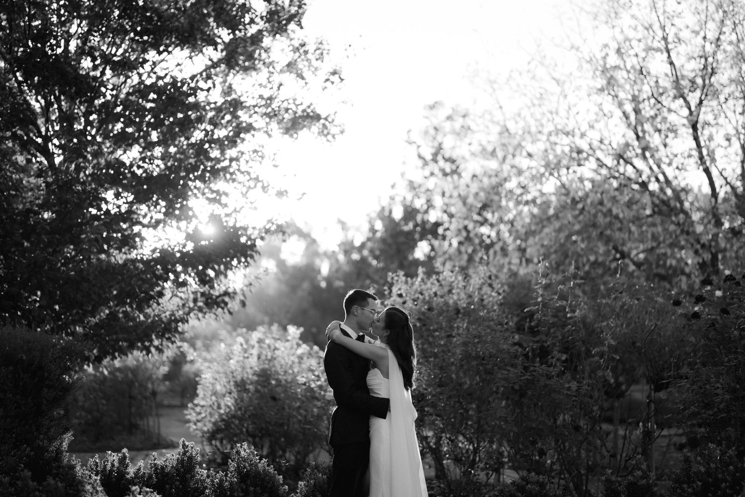 Black and white photo of a bride and groom sharing a romantic kiss surrounded by lush trees in a garden setting.