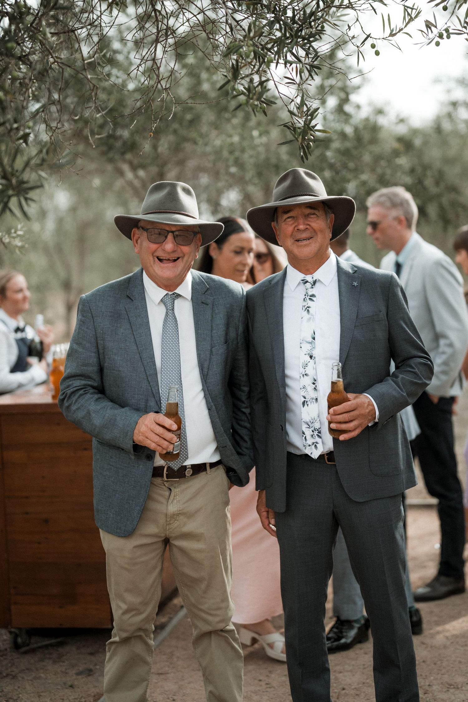 Two men in hats and suits enjoying drinks at an outdoor gathering with olive trees in the background.