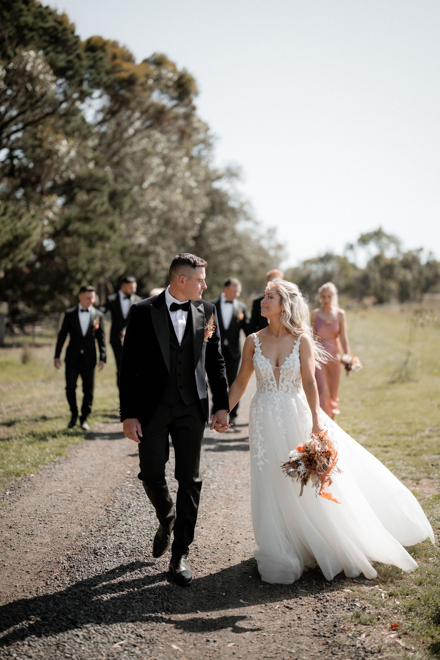 Newlyweds lead wedding party down rural path on sunny day.