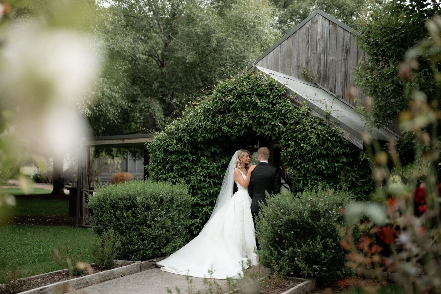 A romantic wedding couple embraces on a garden path next to a rustic wooden structure in a lush green setting.