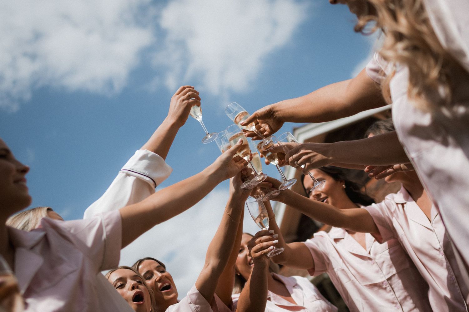 A group of people wearing white outfits raise their hands together joyfully against a bright blue sky with clouds.