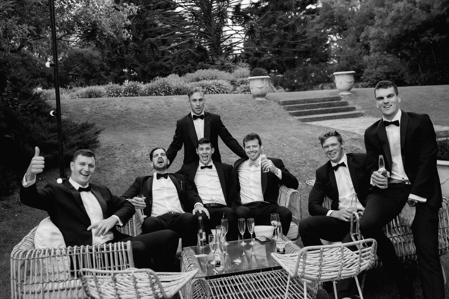 A black and white photograph of people in formal tuxedos relaxing outdoors on patio furniture at a garden gathering.