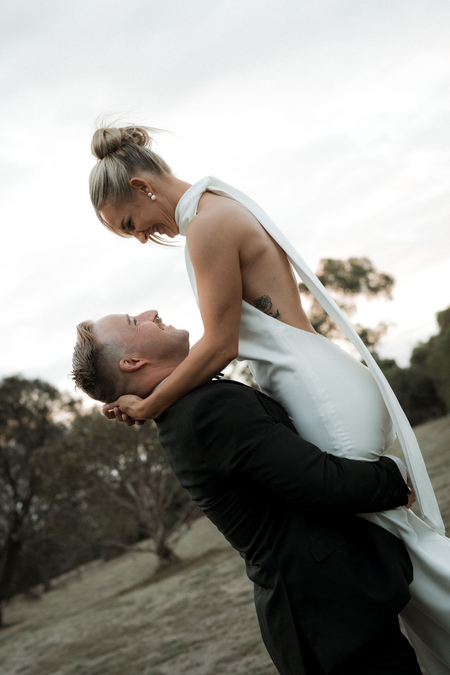 Romantic moment between newlyweds as bride leans over groom against sunset sky.