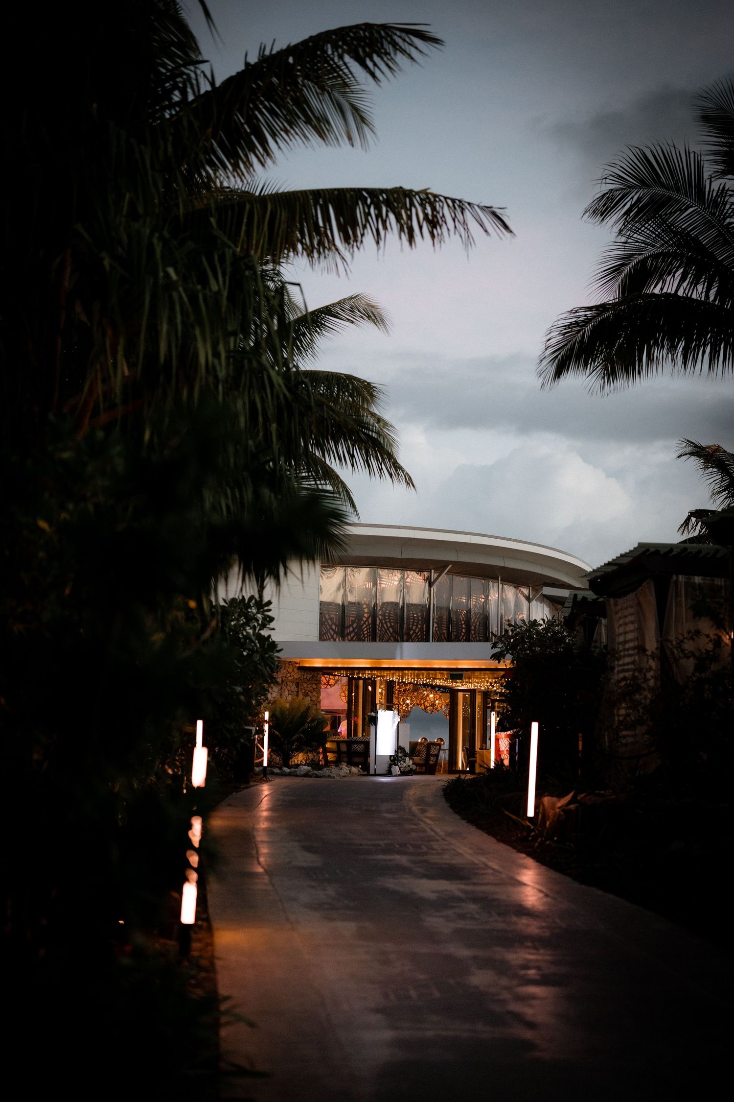 Tropical resort entrance lit at dusk with palm trees and illuminated pathway leading to modern building.
