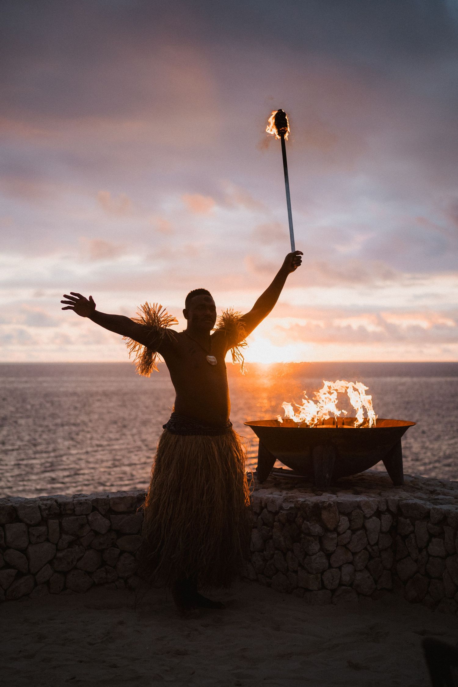 Silhouetted figure in grass skirt performs traditional fire dance at sunset by ocean with flaming torch and fire pit.