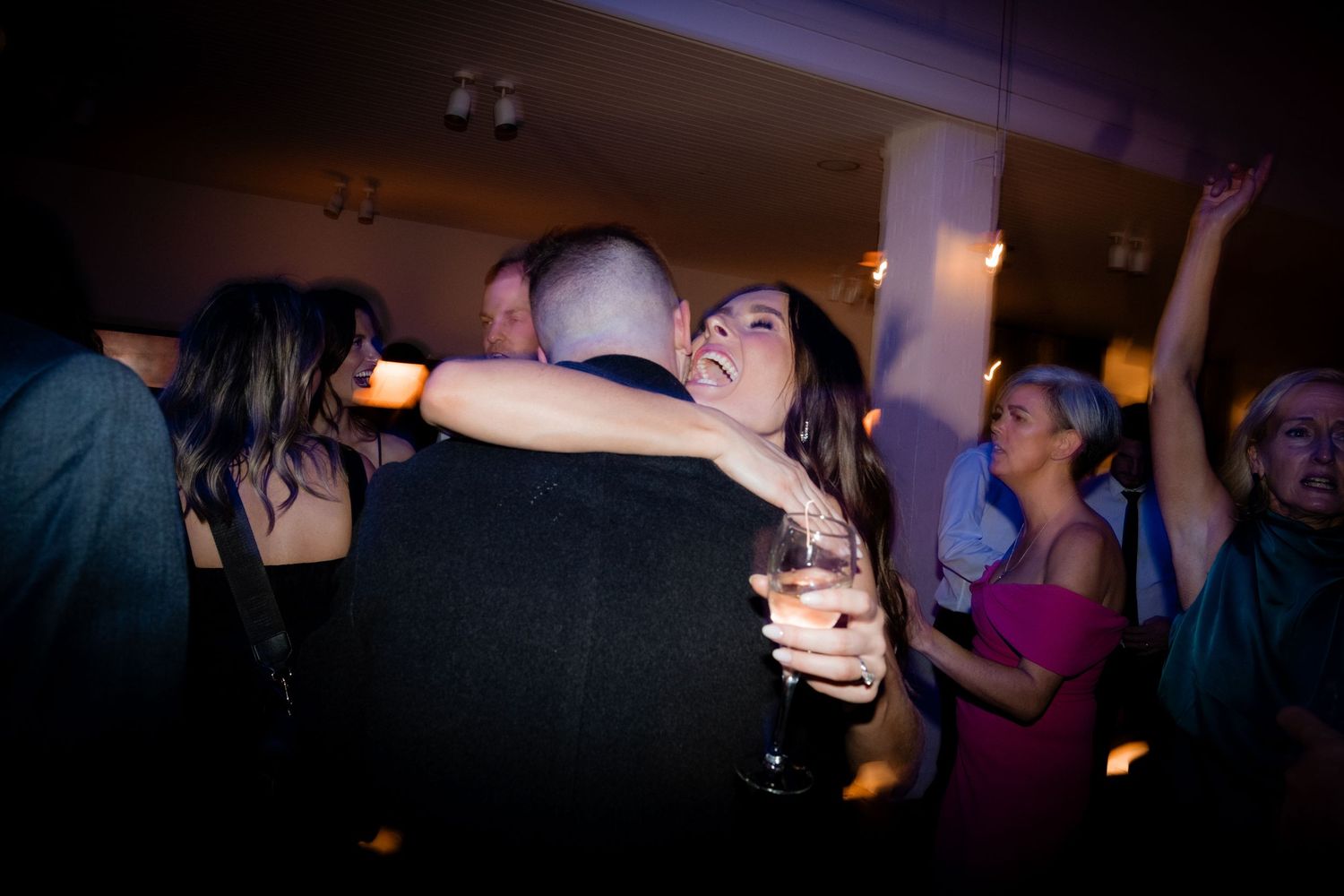 Wedding reception dance floor celebration with guests dancing and embracing in a dimly lit room.