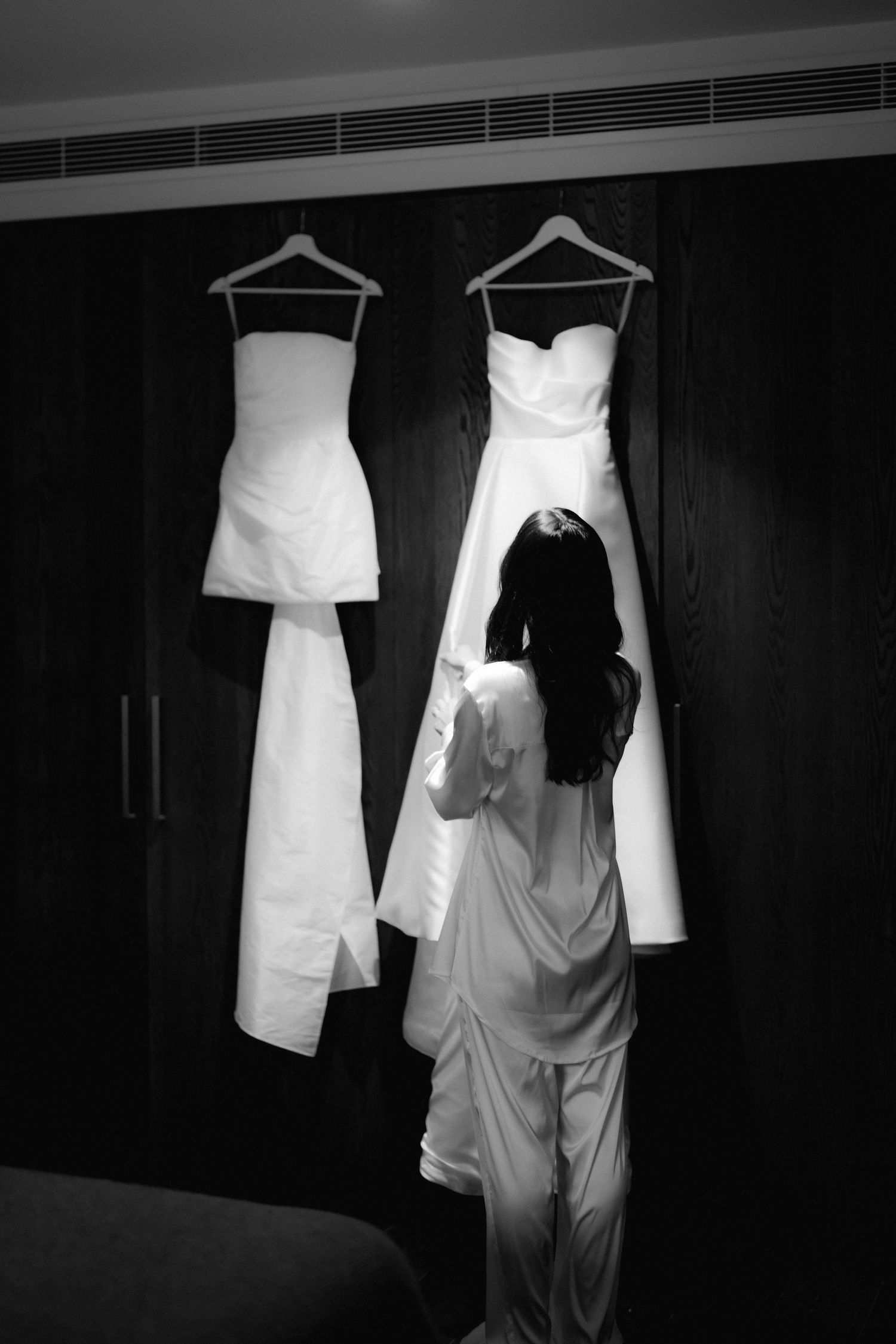 Black and white image of person looking at wedding dresses hanging in wardrobe.