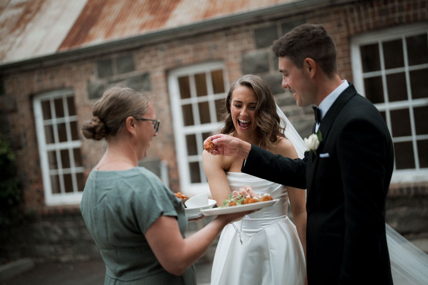 Wedding party celebrating with cake outside a rustic brick building.