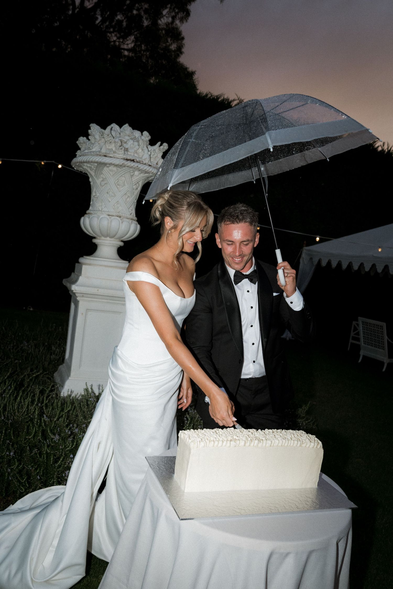Couple cutting their white wedding cake under clear umbrellas at an outdoor reception.