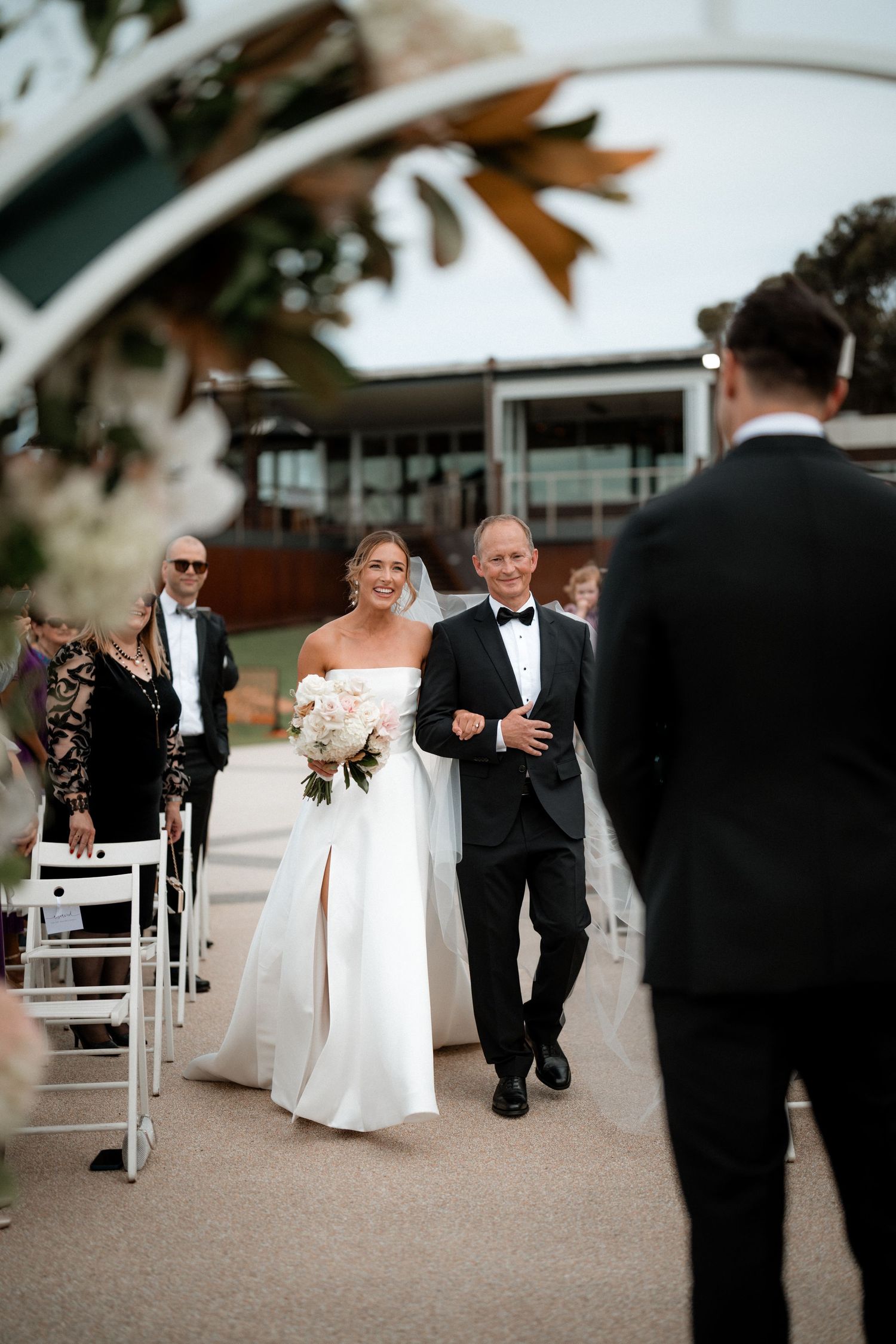 Two figures walk down an outdoor wedding aisle as guests look on during an elegant outdoor ceremony.