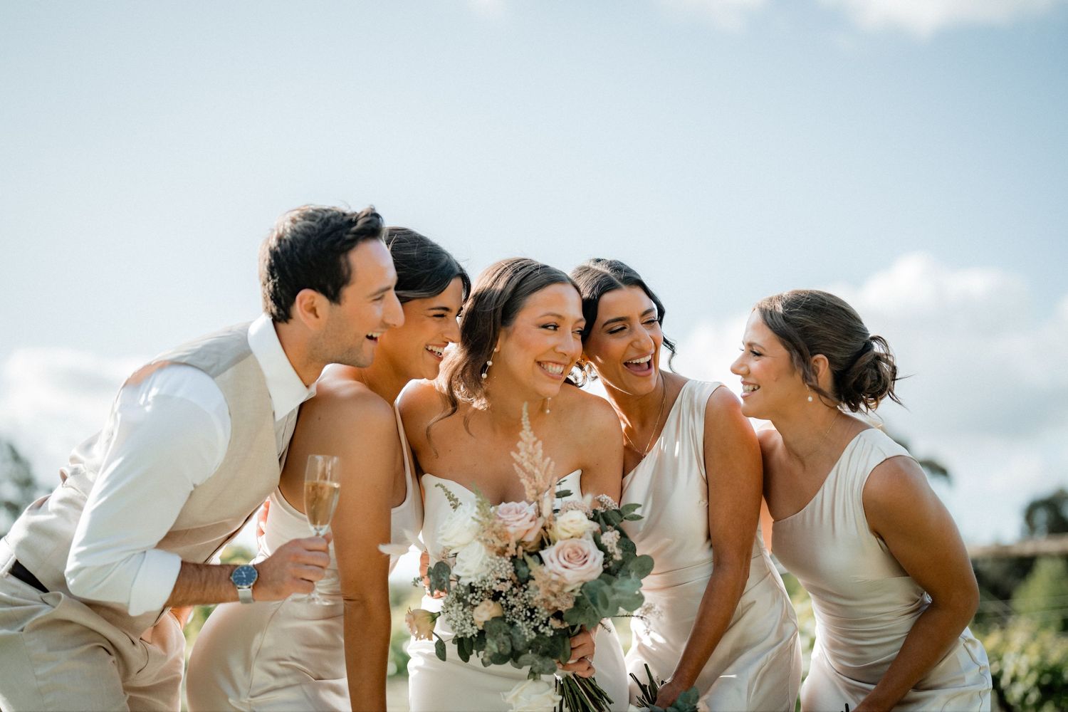 Happy wedding party stands together laughing and embracing outdoors in romantic springtime setting.