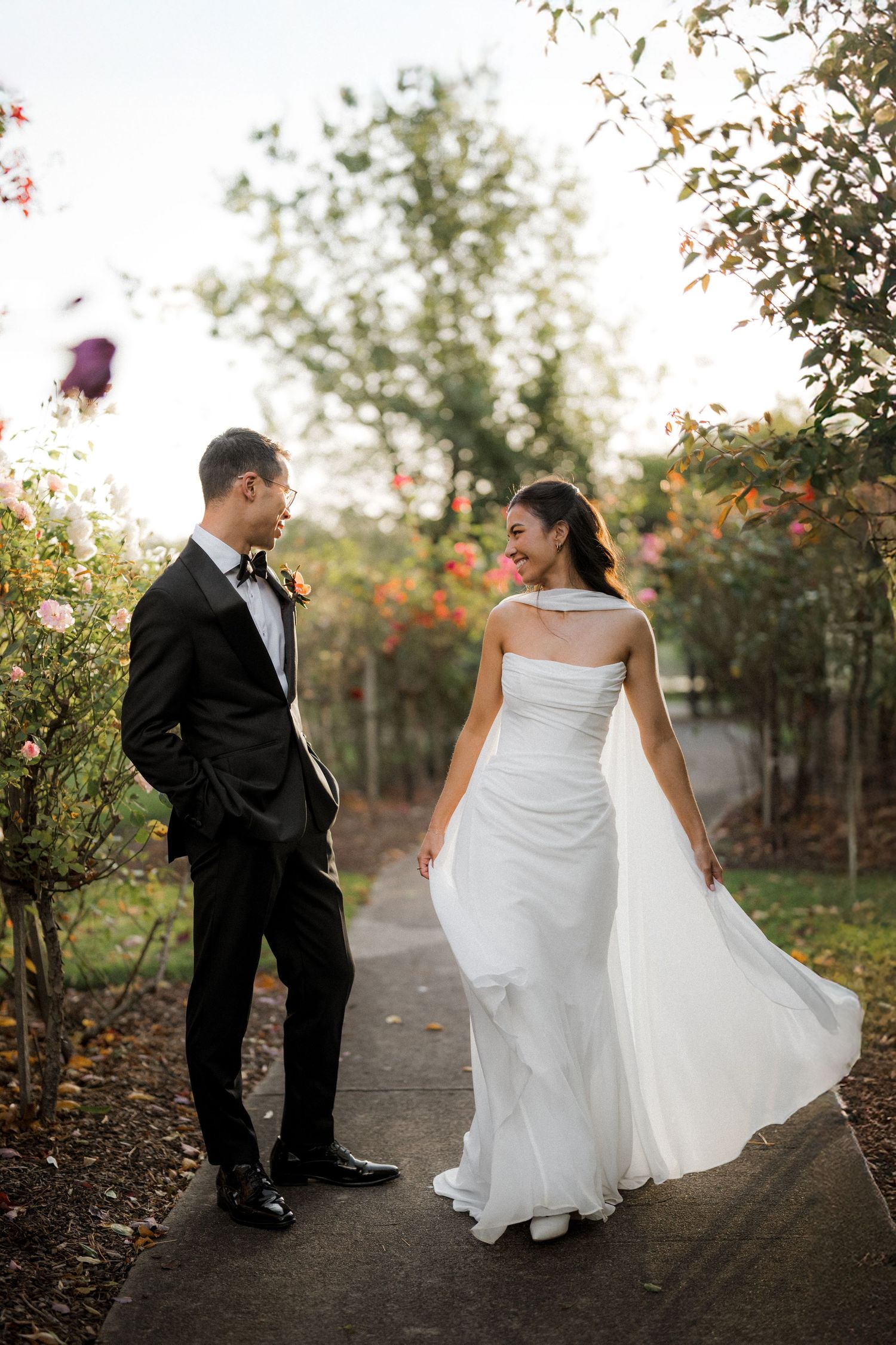 Bride in flowing white gown smiles at groom in black tuxedo on garden path surrounded by roses.