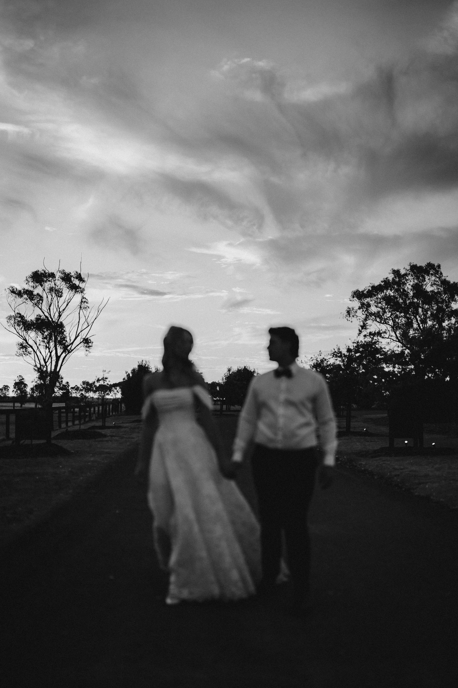 Silhouette of bride and groom holding hands walking at dusk under dramatic sky.