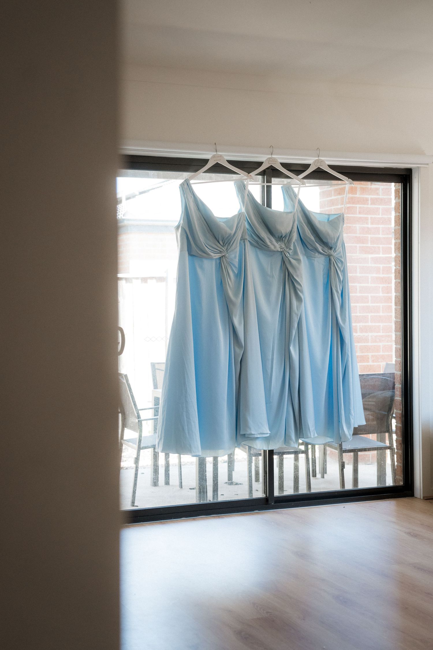 Three light blue bridesmaid dresses hanging on white hangers in front of a sliding glass door.