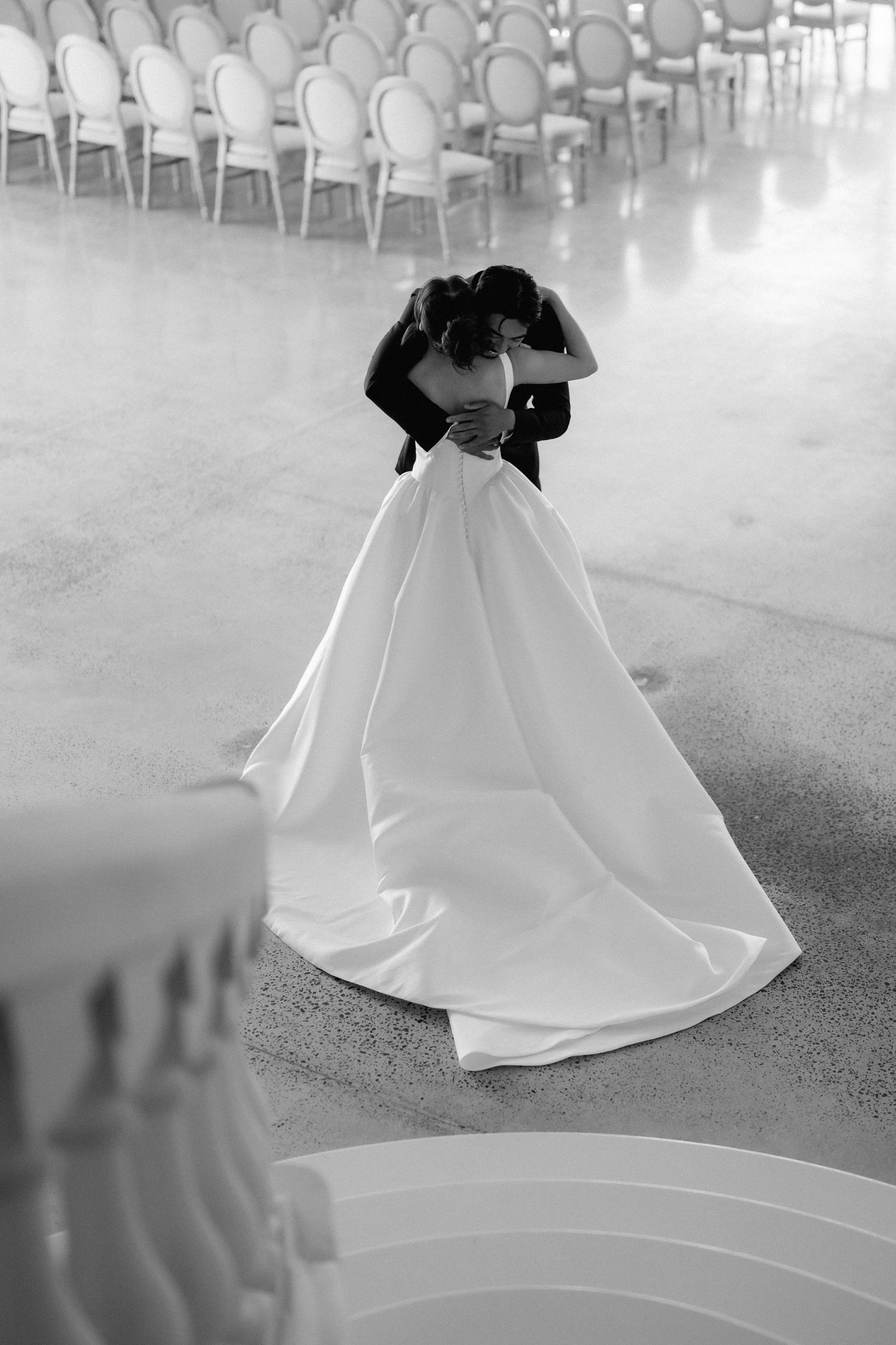 Black and white photo of a bride in a flowing satin gown embracing her groom in an elegant venue with rows of chairs behind them.