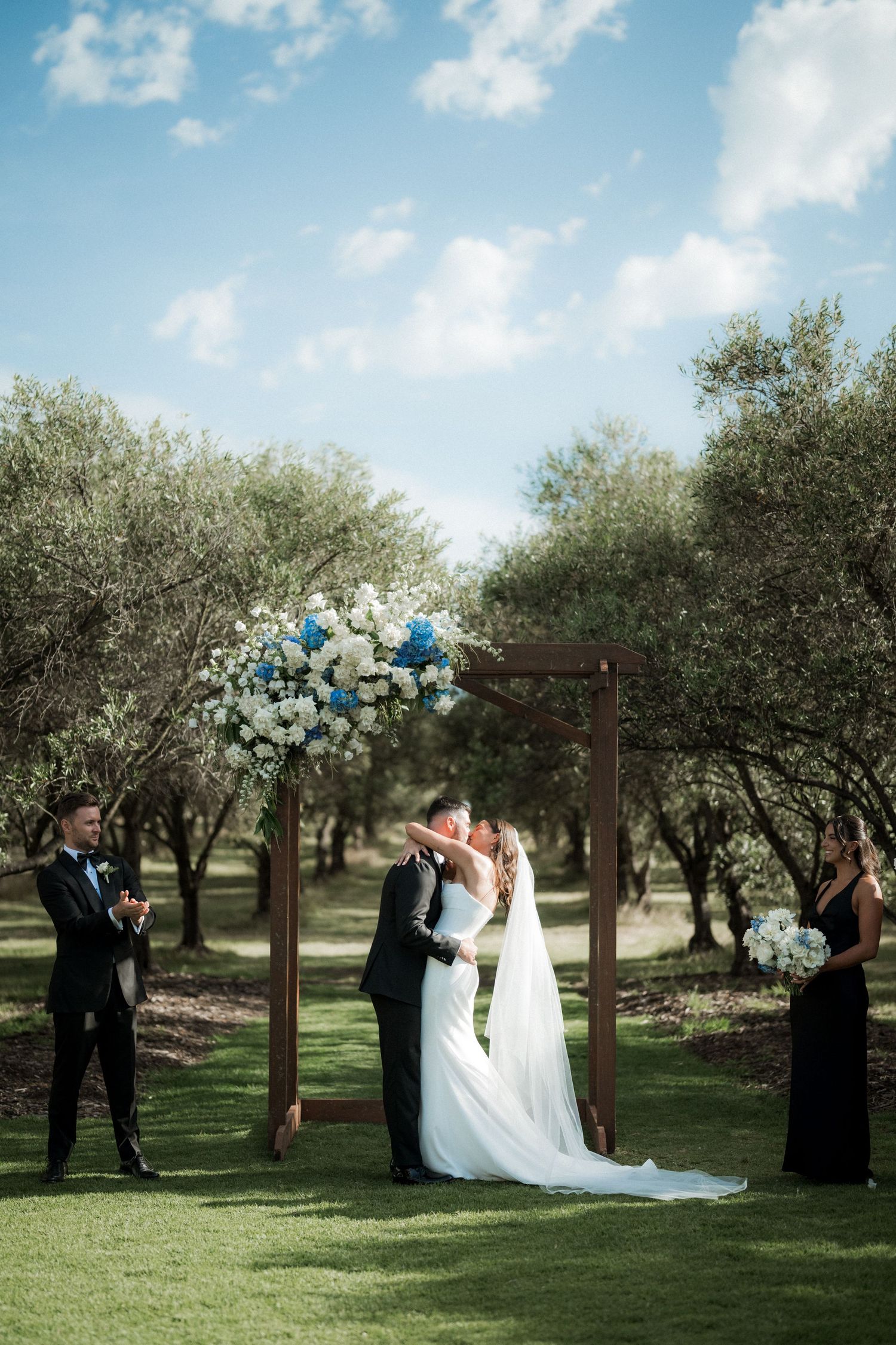 Wedding ceremony setup in an olive grove with a rustic wooden arch decorated in blue and white florals.