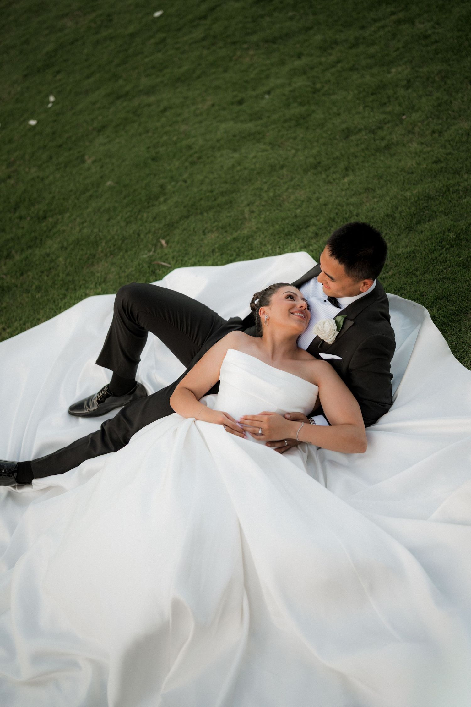 Newlyweds lay together on grass in a romantic moment with the bride's white dress flowing around them.