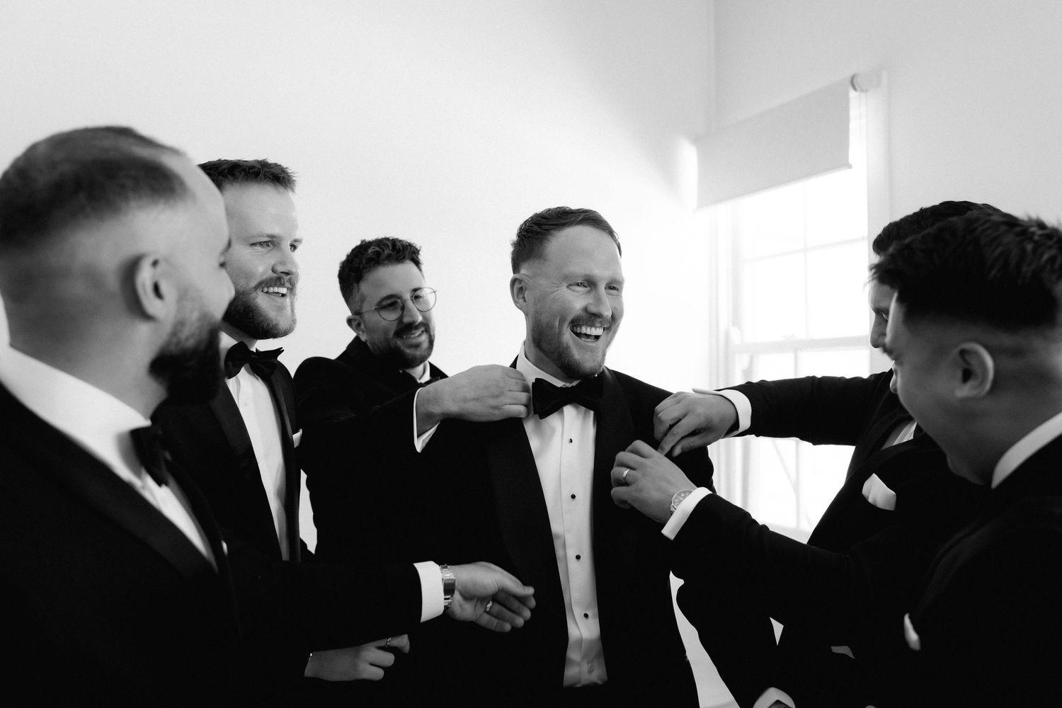 A group of men in formal black suits and ties share a candid laughing moment together at an indoor event.