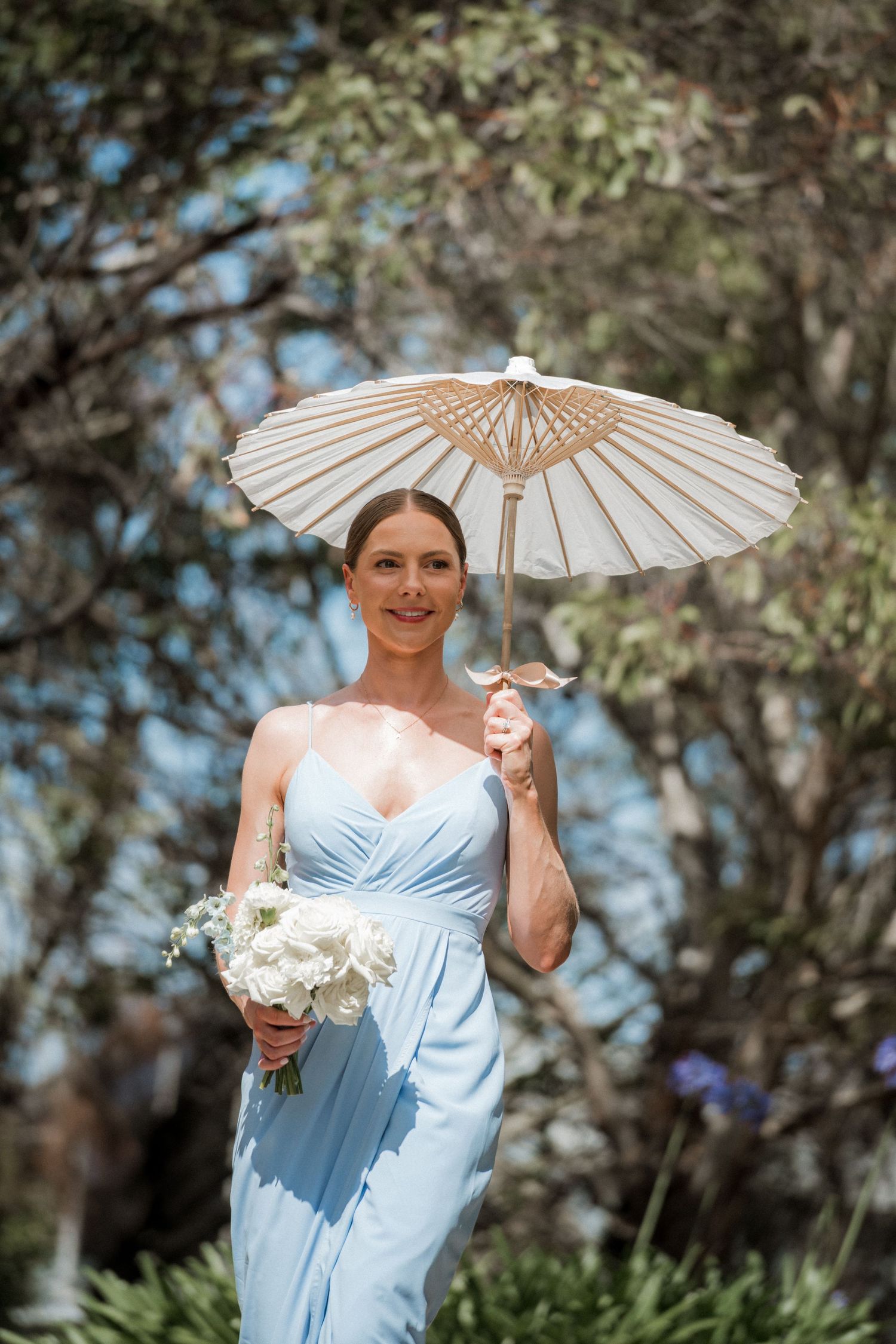 Bridesmaid in light blue dress poses with white parasol in garden setting.