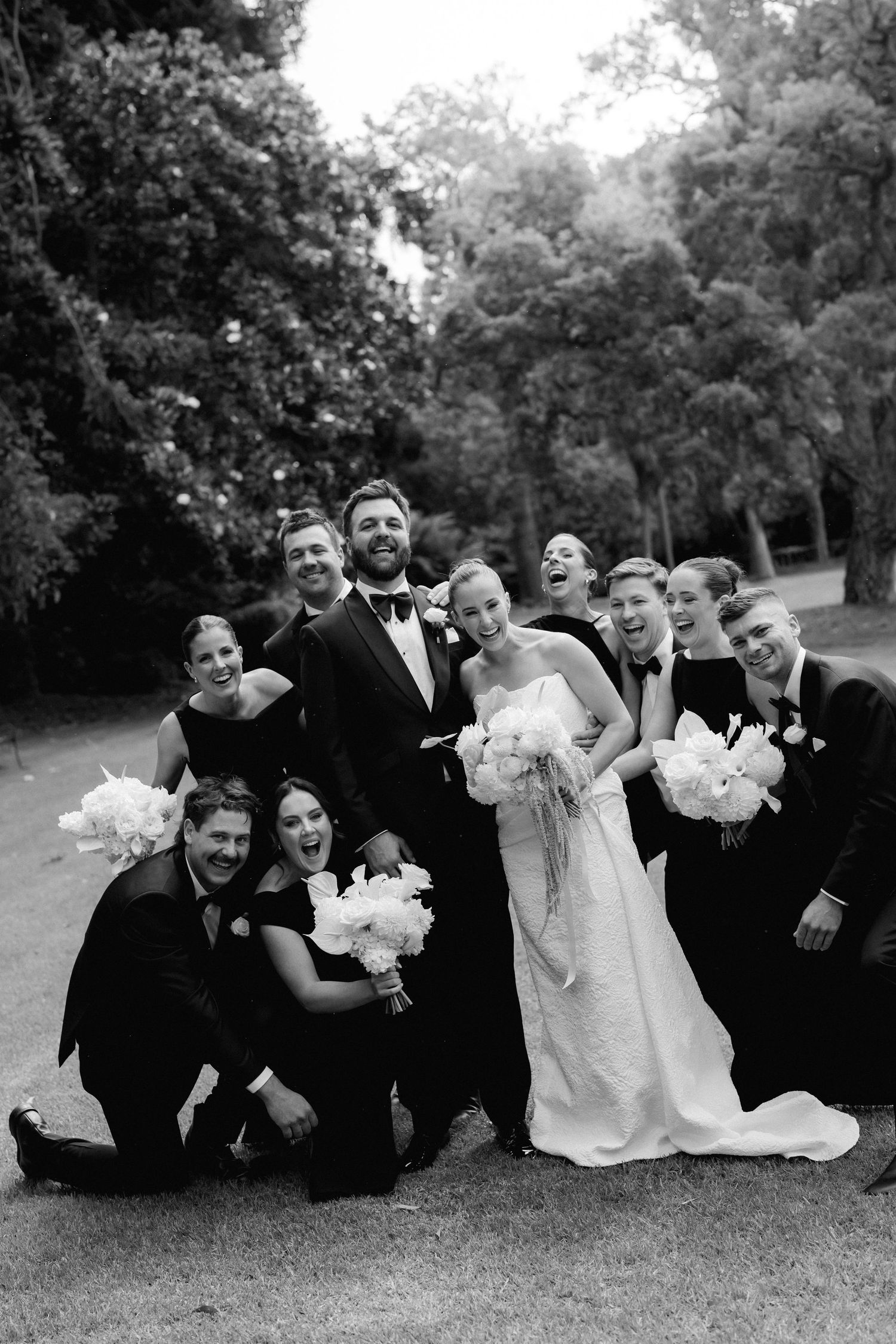A joyful wedding party in black and white formal attire laughs and poses together outdoors among trees.