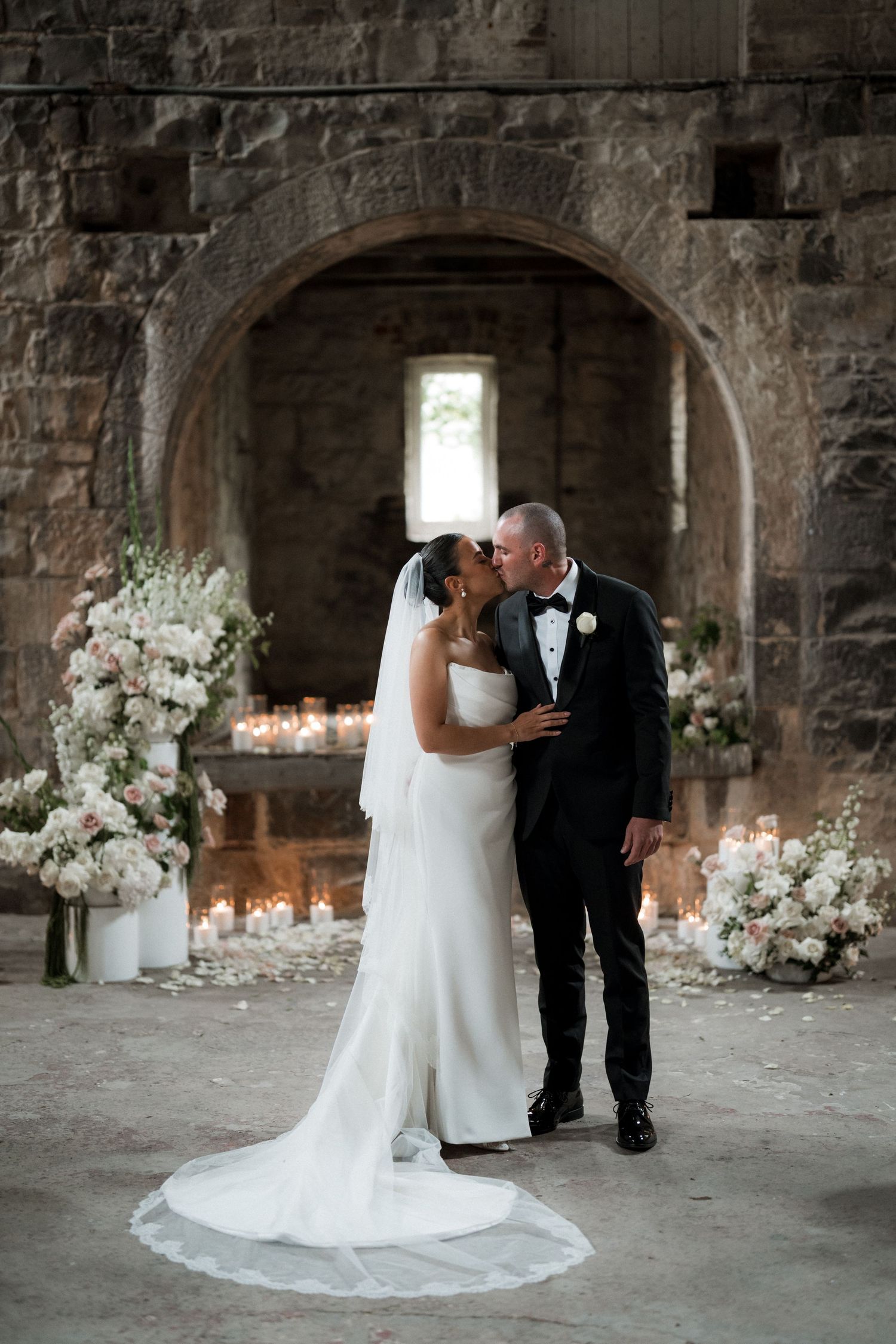 A wedding couple poses in a rustic stone room with elegant white floral arrangements and natural light streaming through windows.