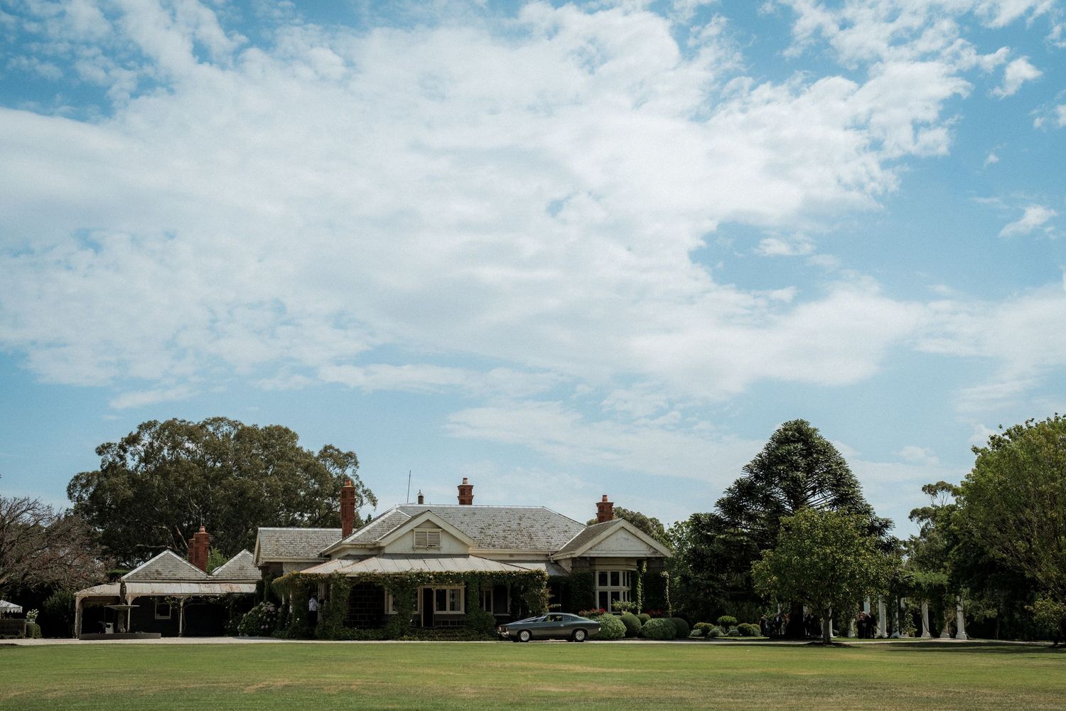 A sprawling country homestead with manicured green lawns, surrounded by tall trees under a bright blue sky.