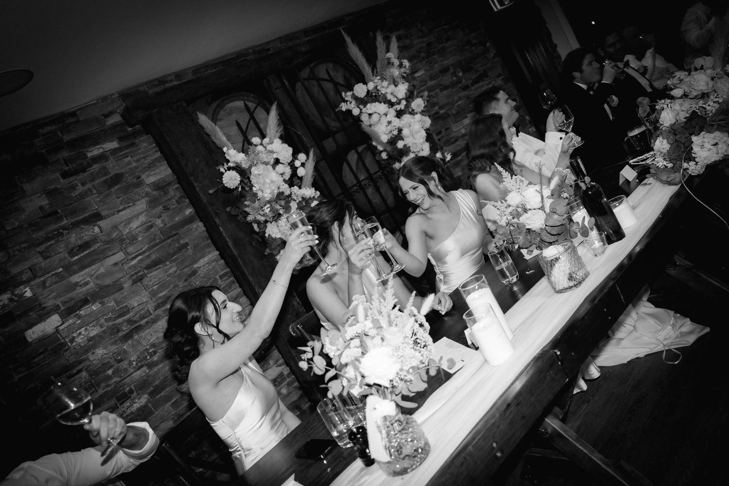 Black and white photo of wedding party celebrating with floral centerpieces on rustic wooden tables.