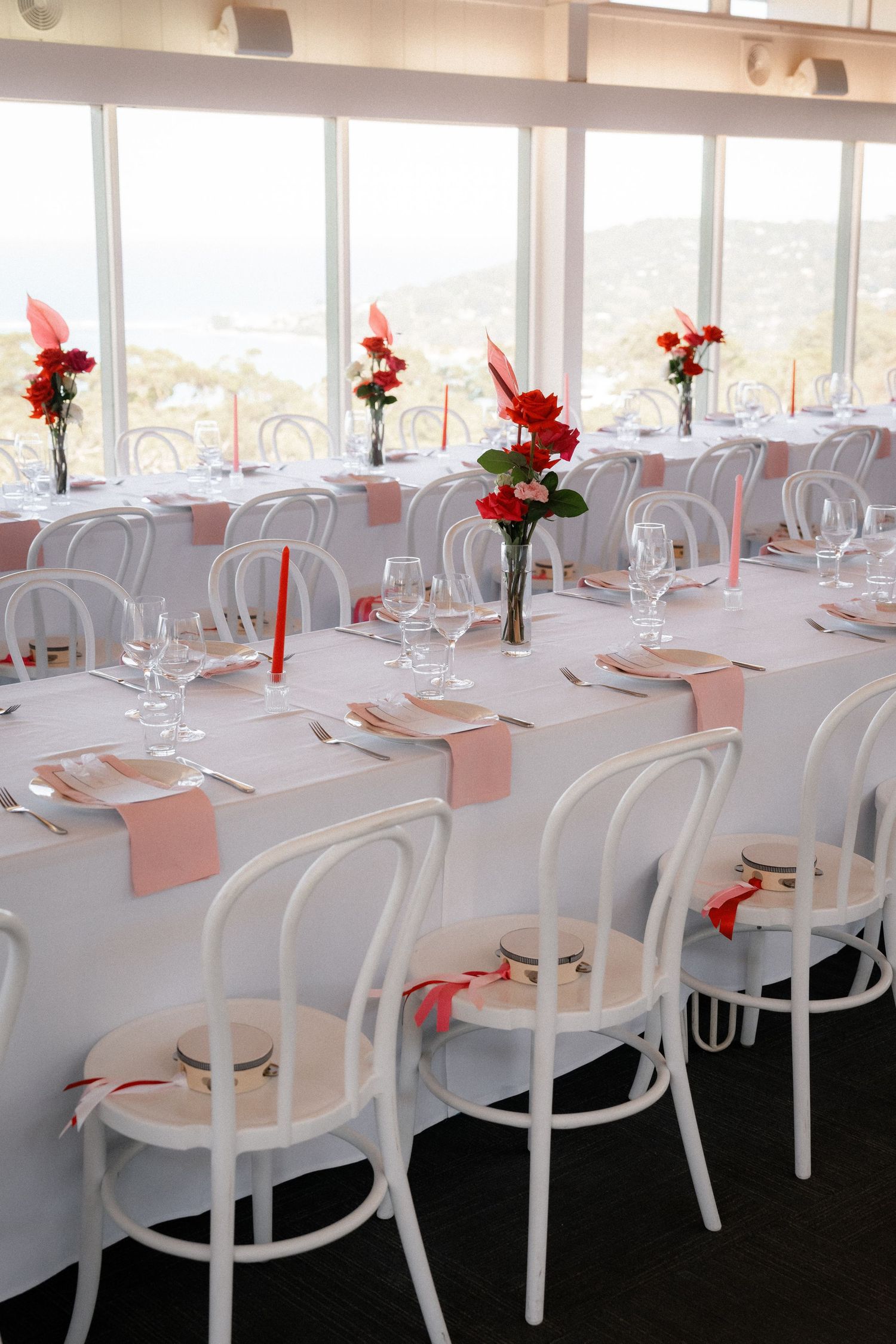 Elegant wedding reception venue with white bentwood chairs, long tables, pink napkins, red floral centerpieces, and ocean views.