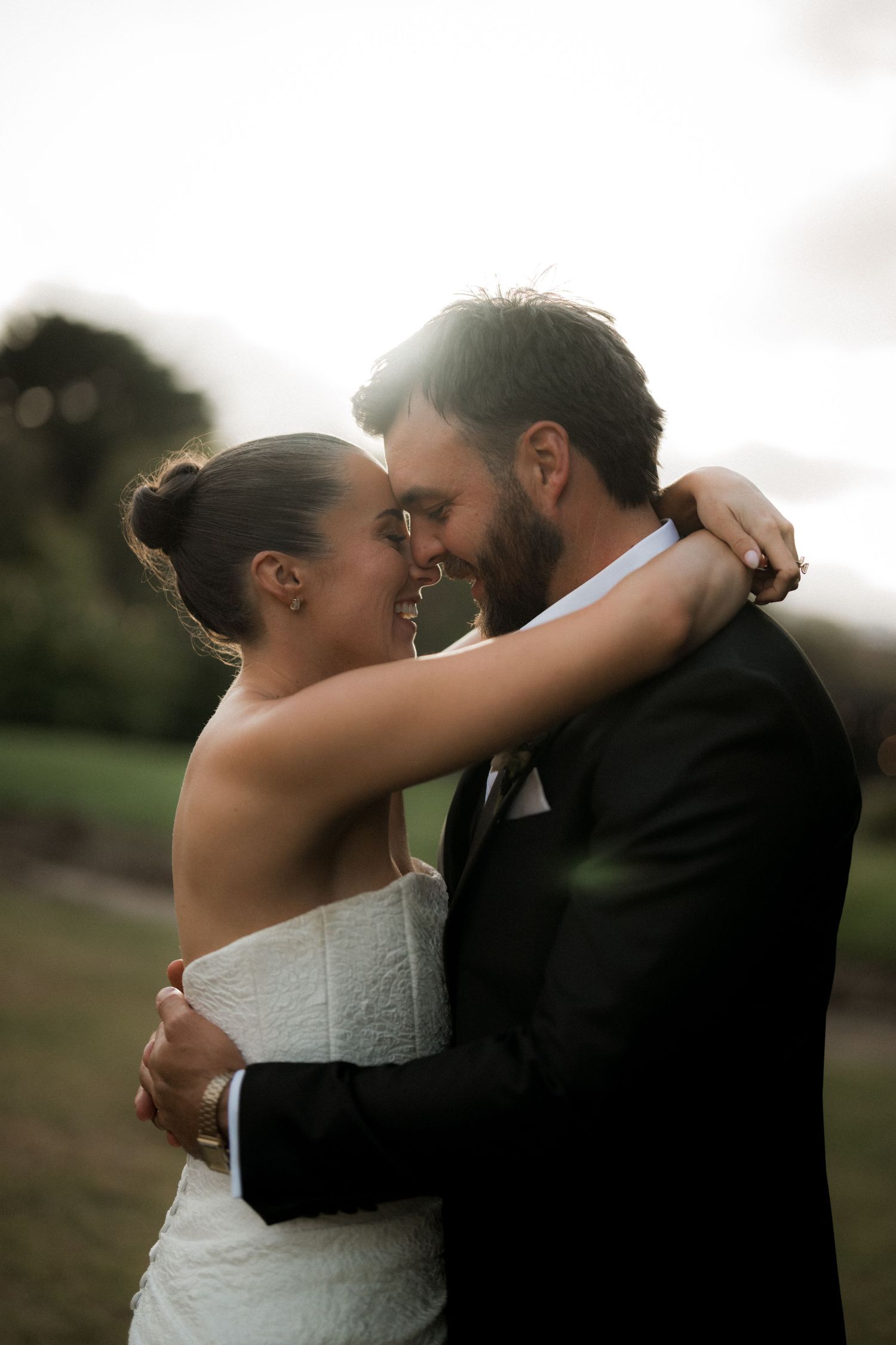Romantic golden hour portrait of bride and groom embracing closely outdoors with warm sunlight glowing behind them.