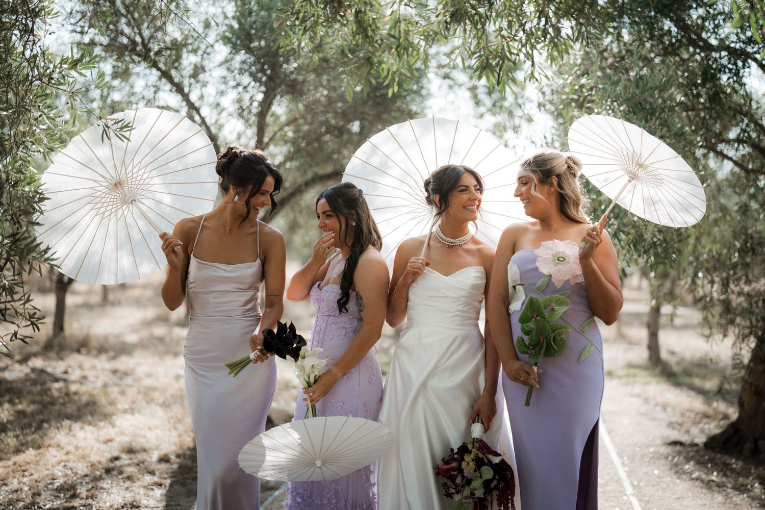 A wedding party in lavender dresses holds white parasols while walking together outdoors on a sunny day.